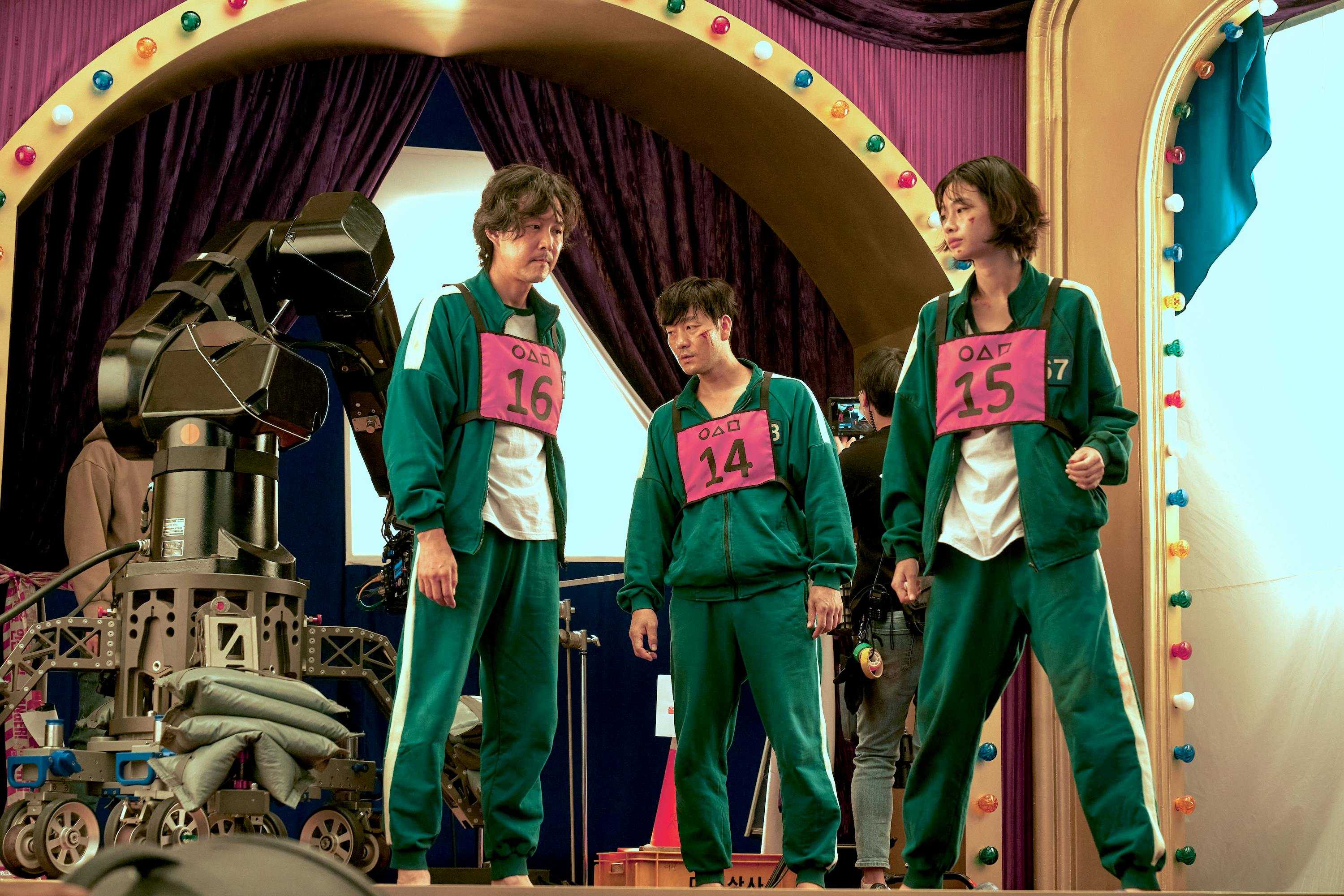 Lee Jung-jae, Park Hae-soo, and Jung Ho-yeon wear their green tracksuits with their numbers in pink on their chest. They are surrounded by colorful lights and camera gear.