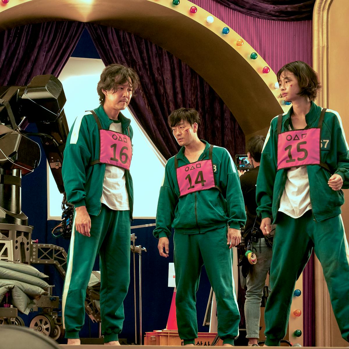 Lee Jung-jae, Park Hae-soo, and Jung Ho-yeon wear their green tracksuits with their numbers in pink on their chest. They are surrounded by colorful lights and camera gear.