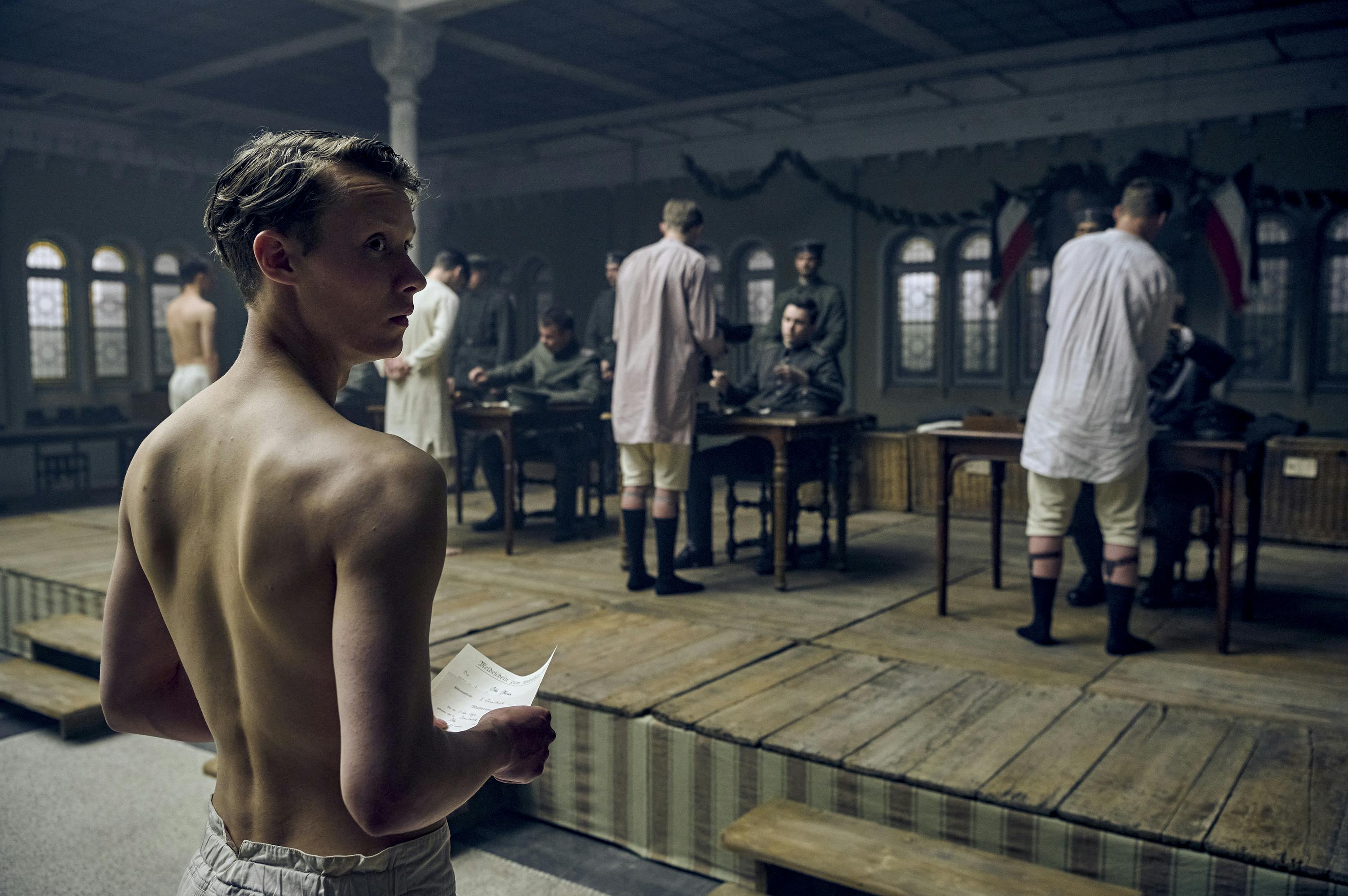 Paul Bäumer (Felix Kammerer) stands in white shorts waiting his turn to receive his uniform. In front of him are other young men in white undergarments accepting their uniforms from men at desks.