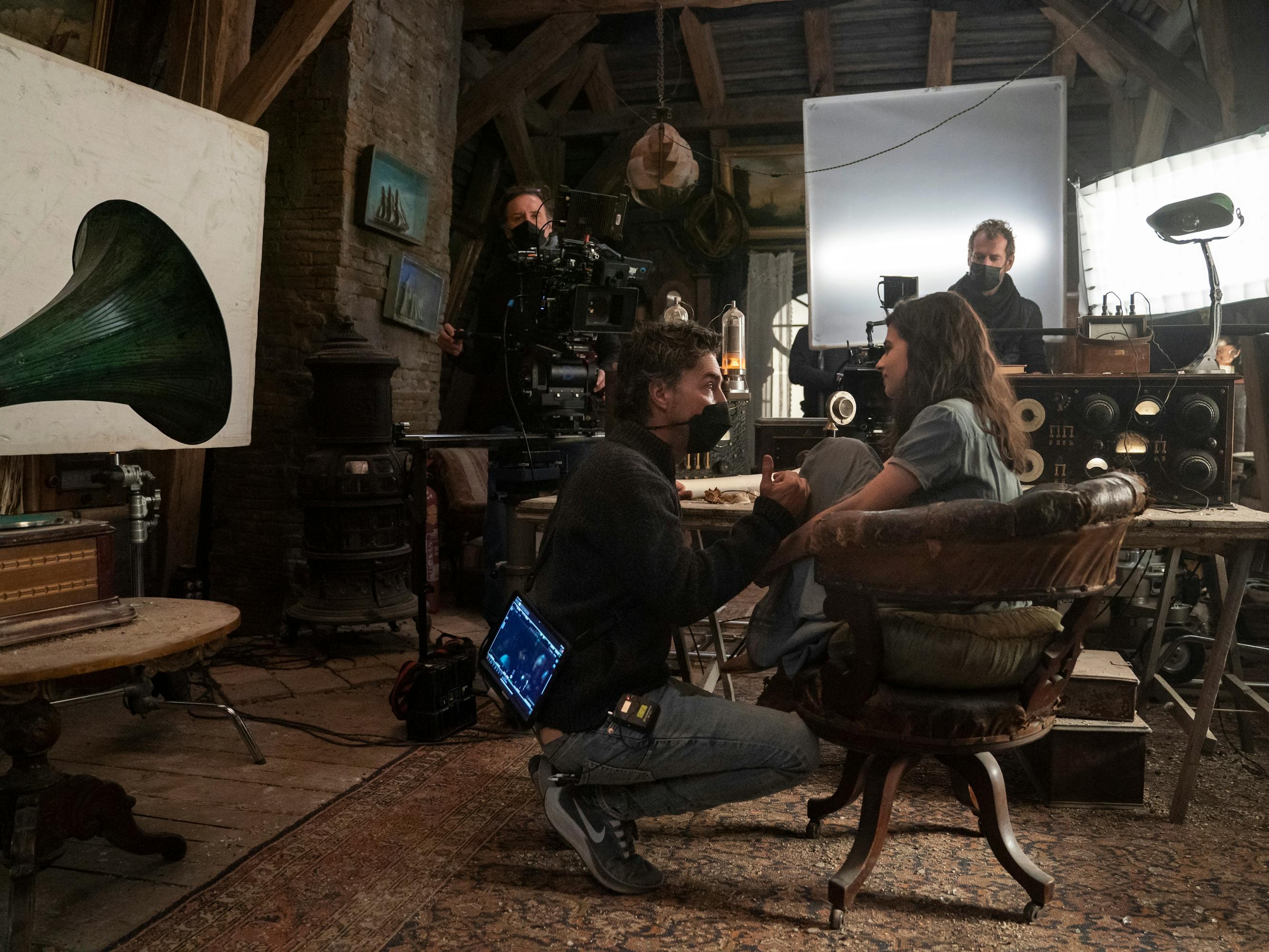 Shawn Levy and Aria Mia Loberti behind the scenes on set. Loberti sits at a desk in a large leather chair and Levy wears a mask and kneels to her level.