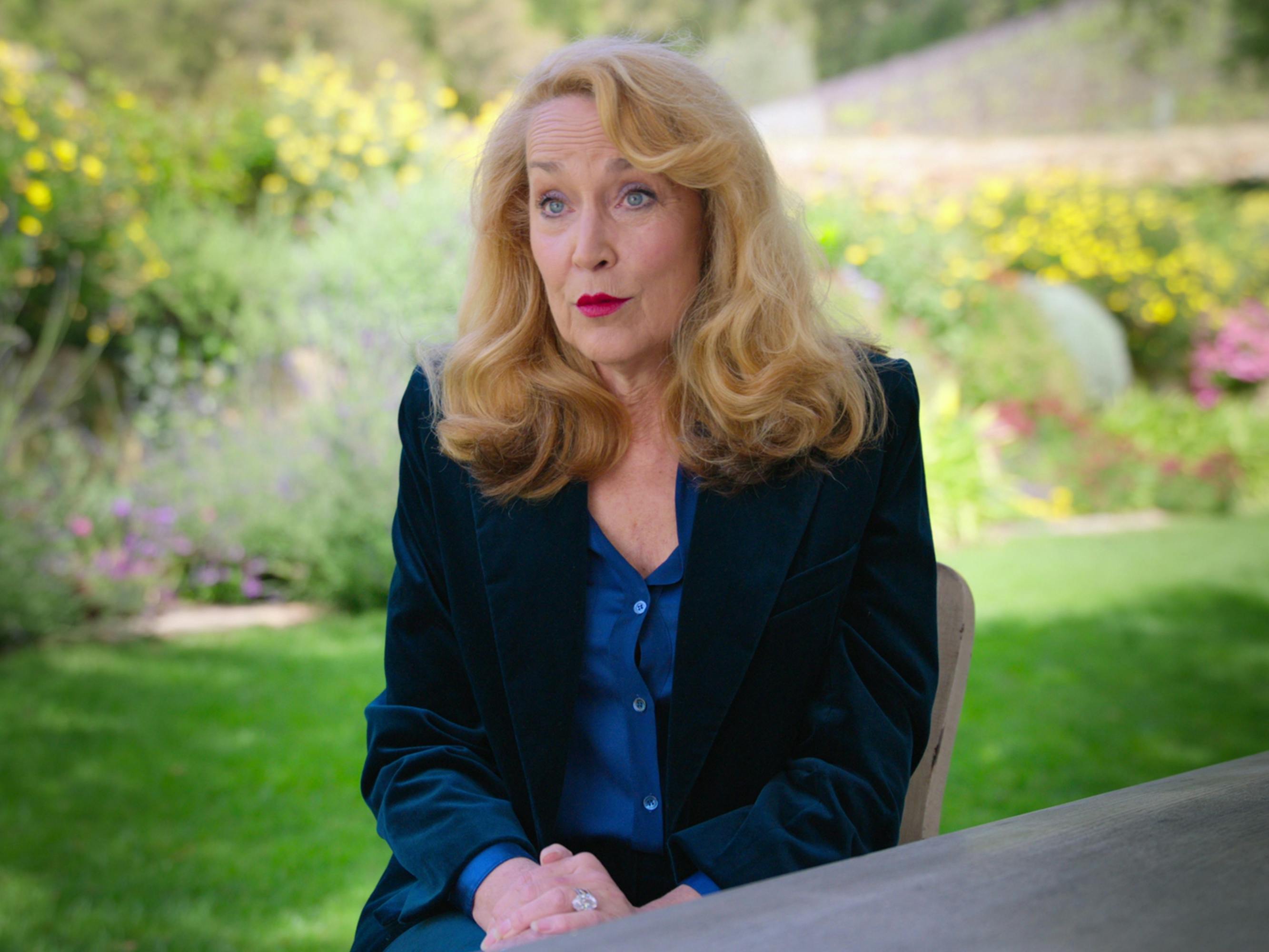 Jerry Hall wears a blue shirt and dark blazer and a bold red lip.