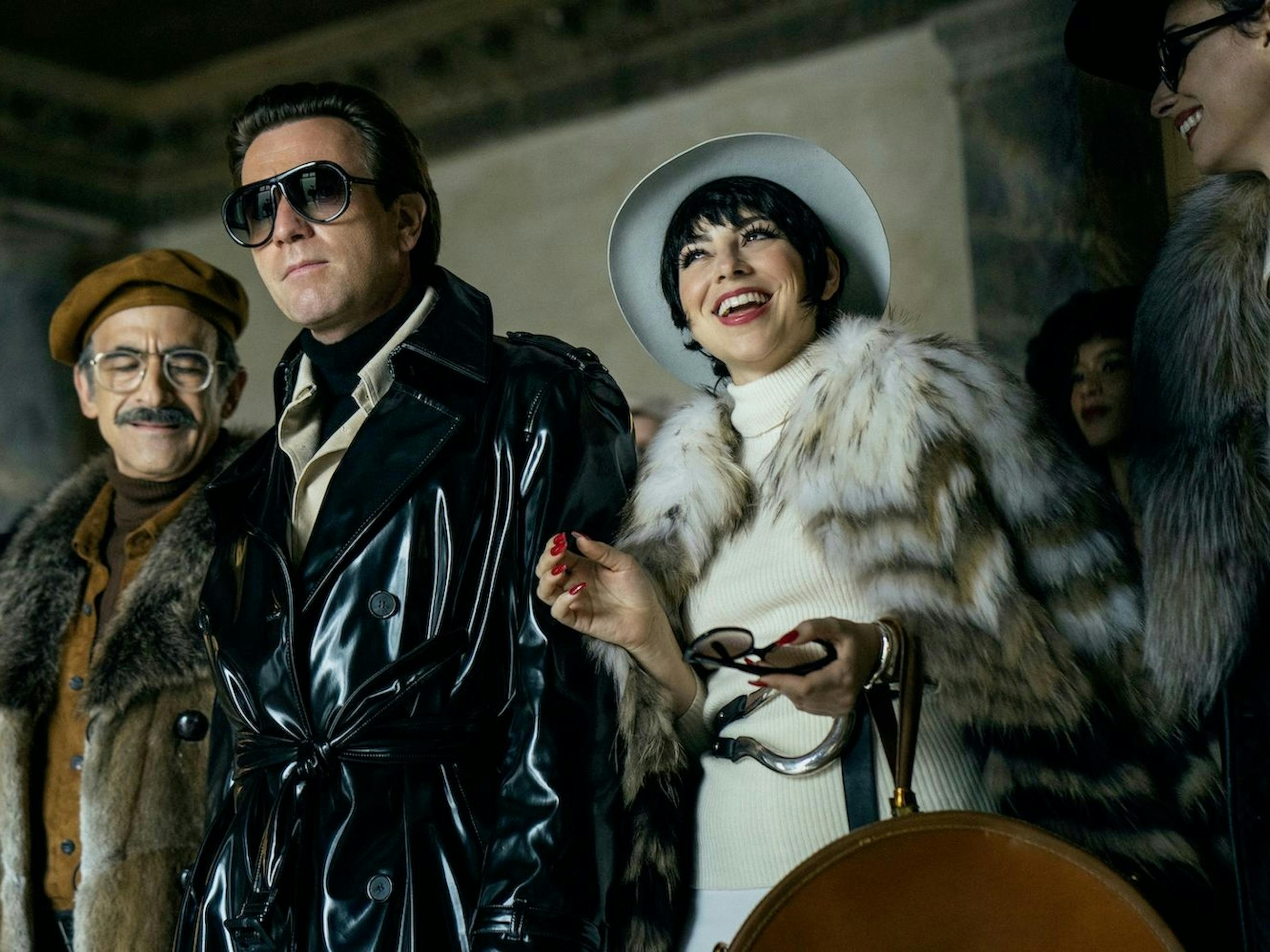 Joe Eula (David Pittu), Halston (Ewan McGregor), and Liza Minnelli (Krysta Rodriguez) stand in a row. Eula wears a fur coat, beret, glasses, and a mustache. Halston wears his signature jacket, and dark sunglasses. Minnelli wears all white, including a hat and fur.