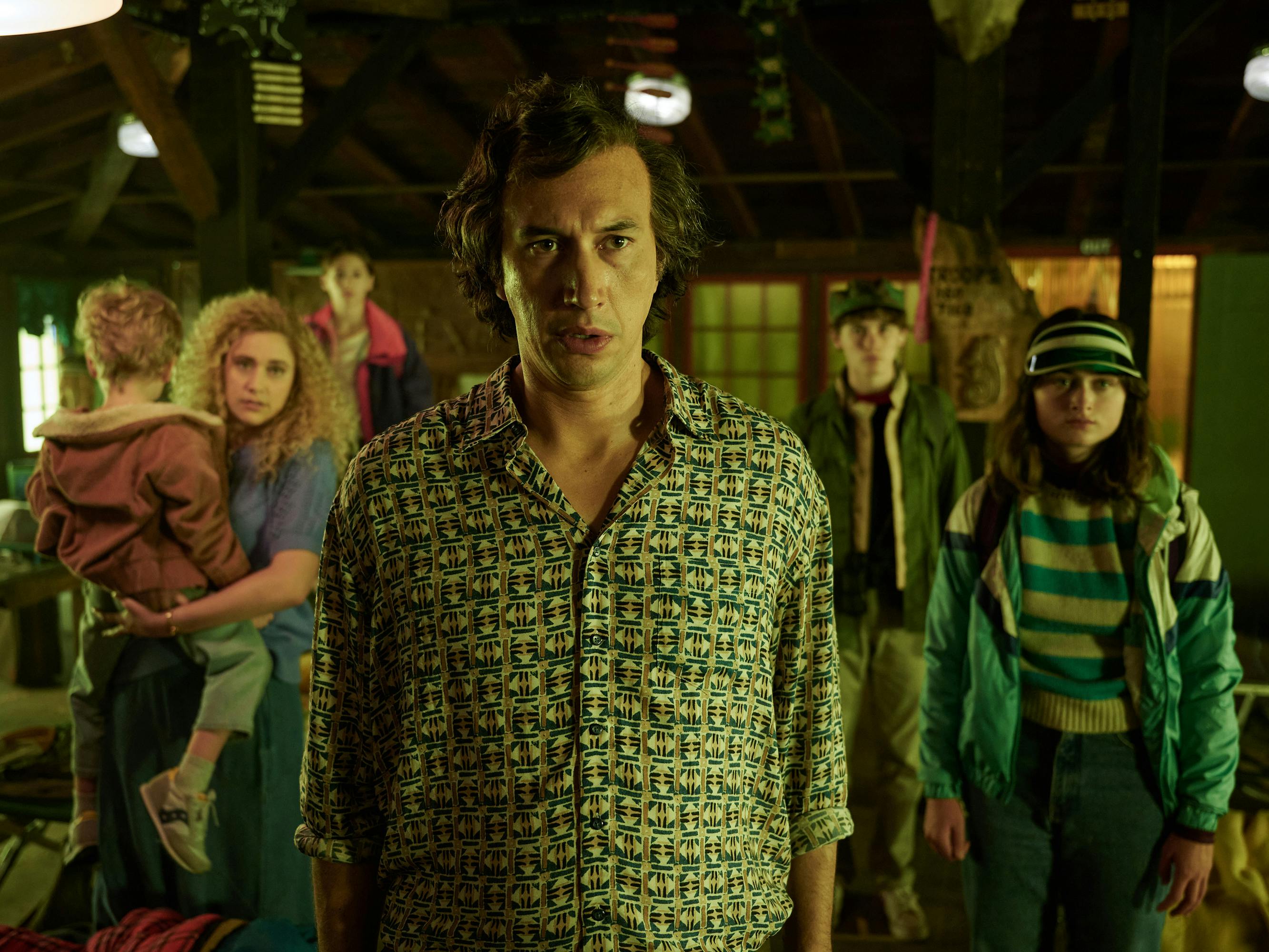 The Gladney family stand facing something offscreen. Greta Gerwig has a big perm and holds a baby, and Adam Driver faces the camera head on in a patterned shirt.