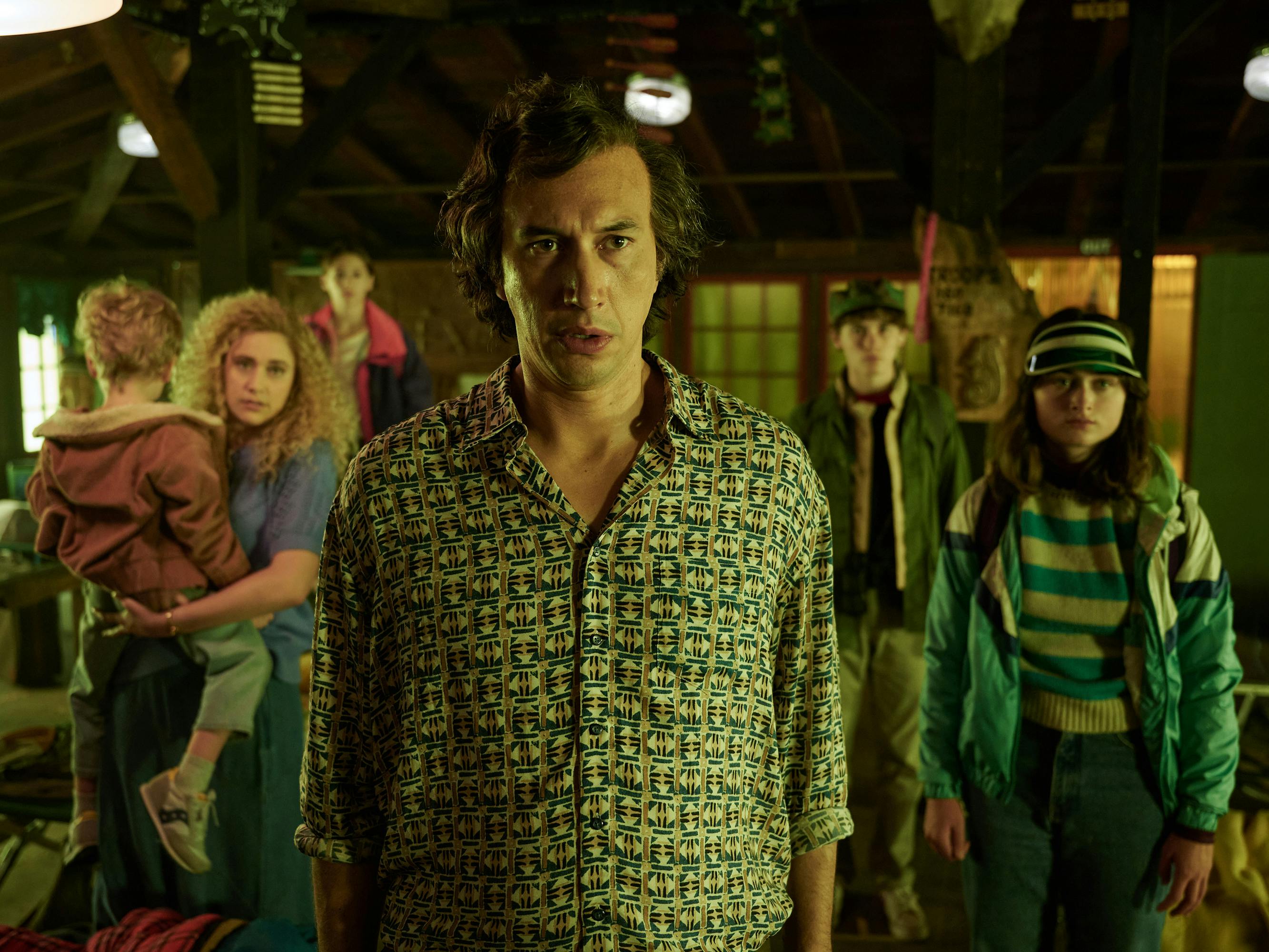 The Gladney family stand facing something offscreen. Greta Gerwig has a big perm and holds a baby, and Adam Driver faces the camera head on in a patterned shirt. 