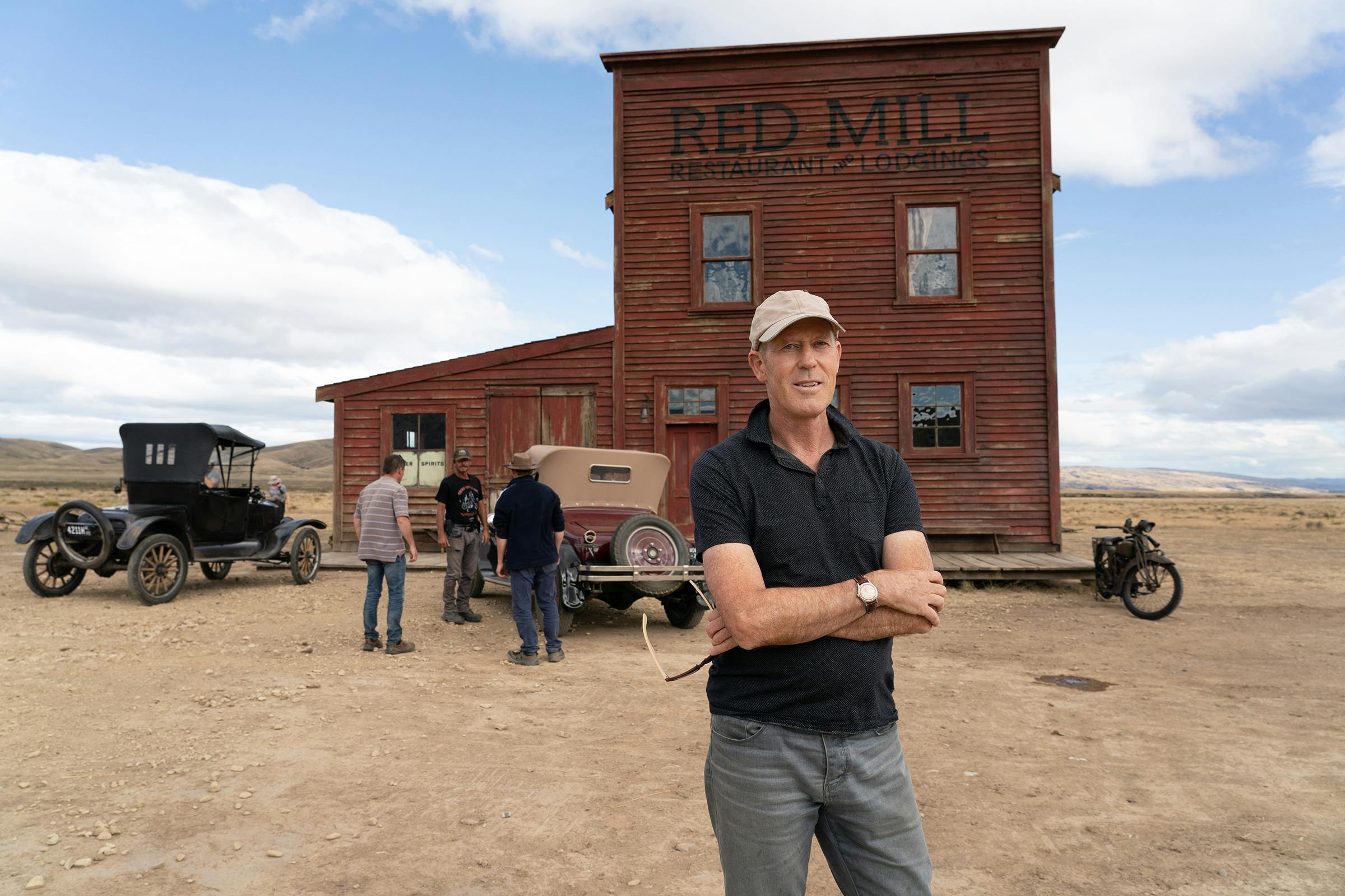 Grant Major stands outside the Red Mill set — which is on wheels — with his arms crossed. The sky is blue with puffy white clouds, the ground is sandy and dusty. Major wears a black polo shirt and grey pants. People and buggies dot the background.