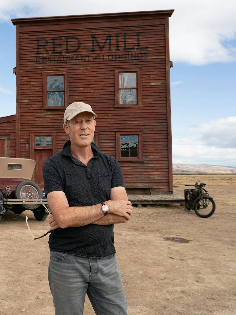 Grant Major stands outside the Red Mill set — which is on wheels — with his arms crossed. The sky is blue with puffy white clouds, the ground is sandy and dusty. Major wears a black polo shirt and grey pants. People and buggies dot the background.