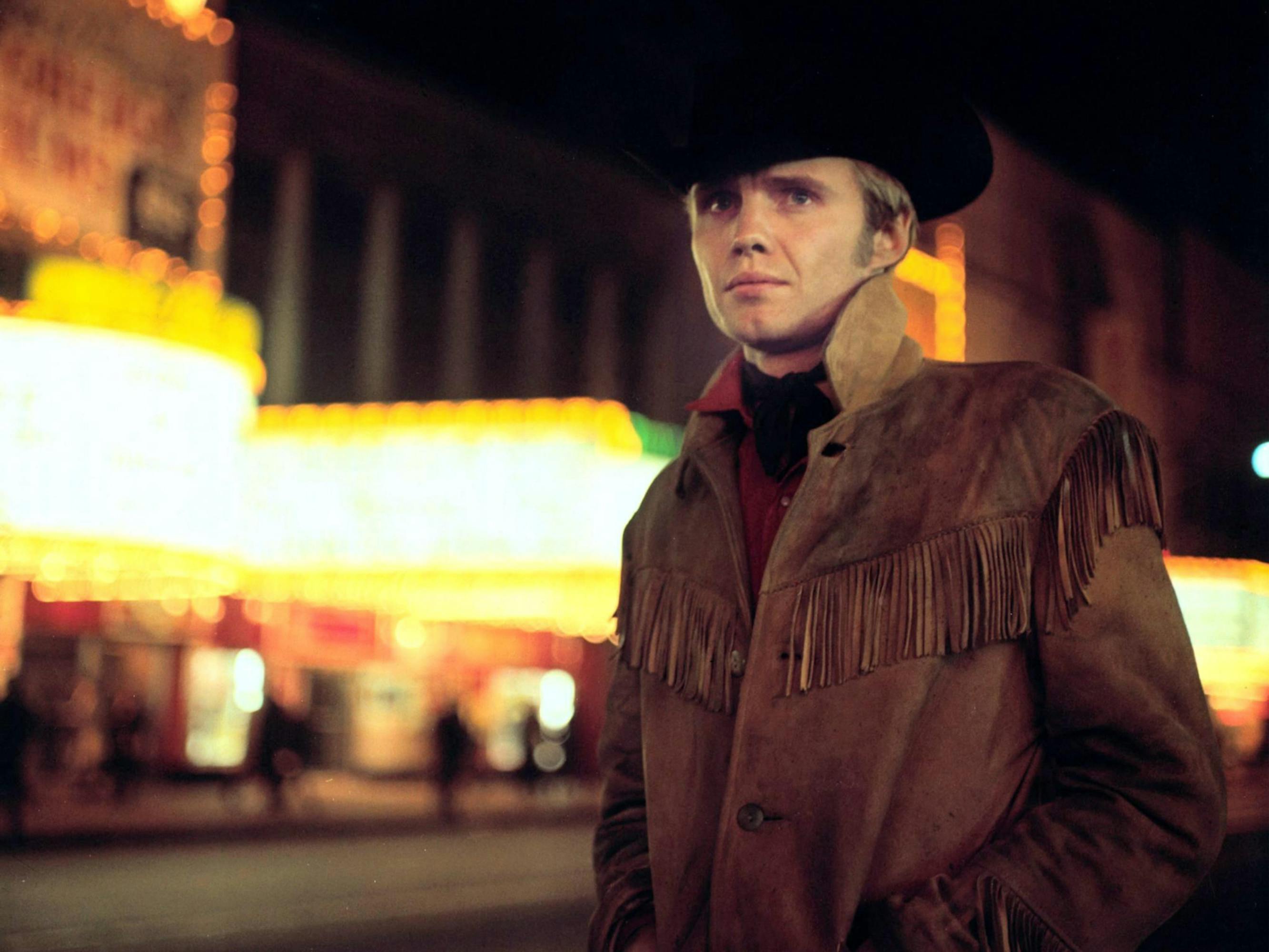 Jon Voight stands in his cowboy hat and jacket, with glowing neon lights blurred behind him.