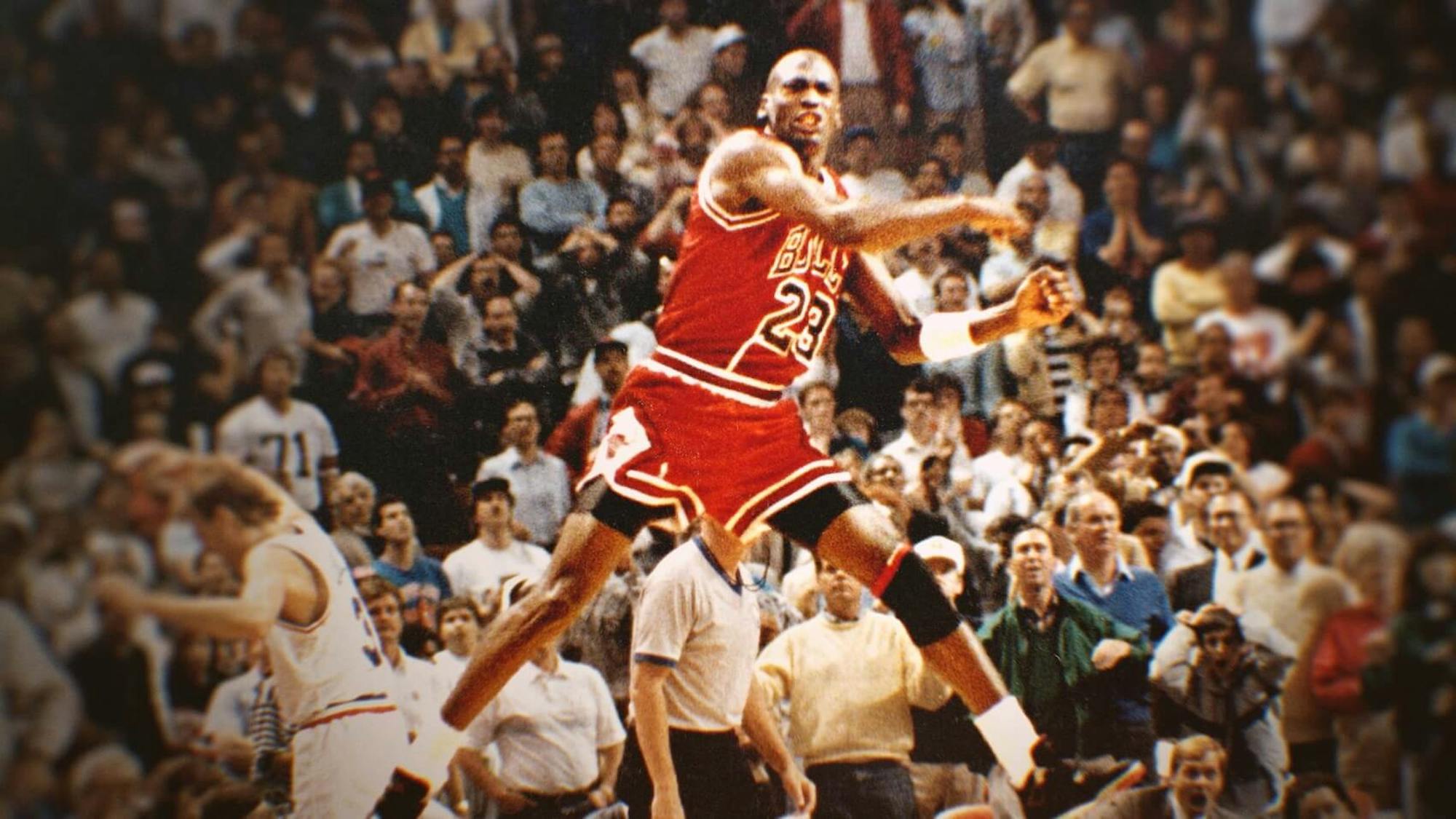 Michael Jordan jumps in the center of this lively shot. The people in the background appear on the edge of their seats. Jordan wears a red uniform with the number 23.