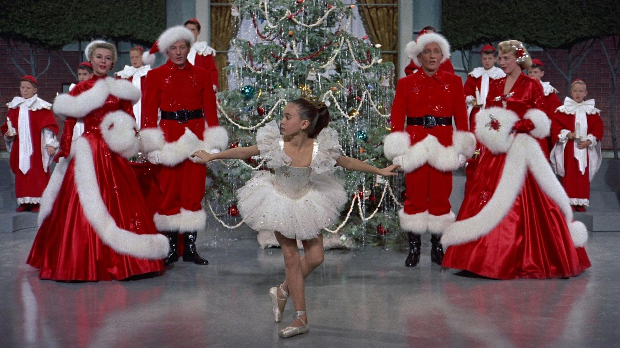 The cast of White Christmas lines up in Santa costumes. In the center is a huge tree and a small girl in a tutu.