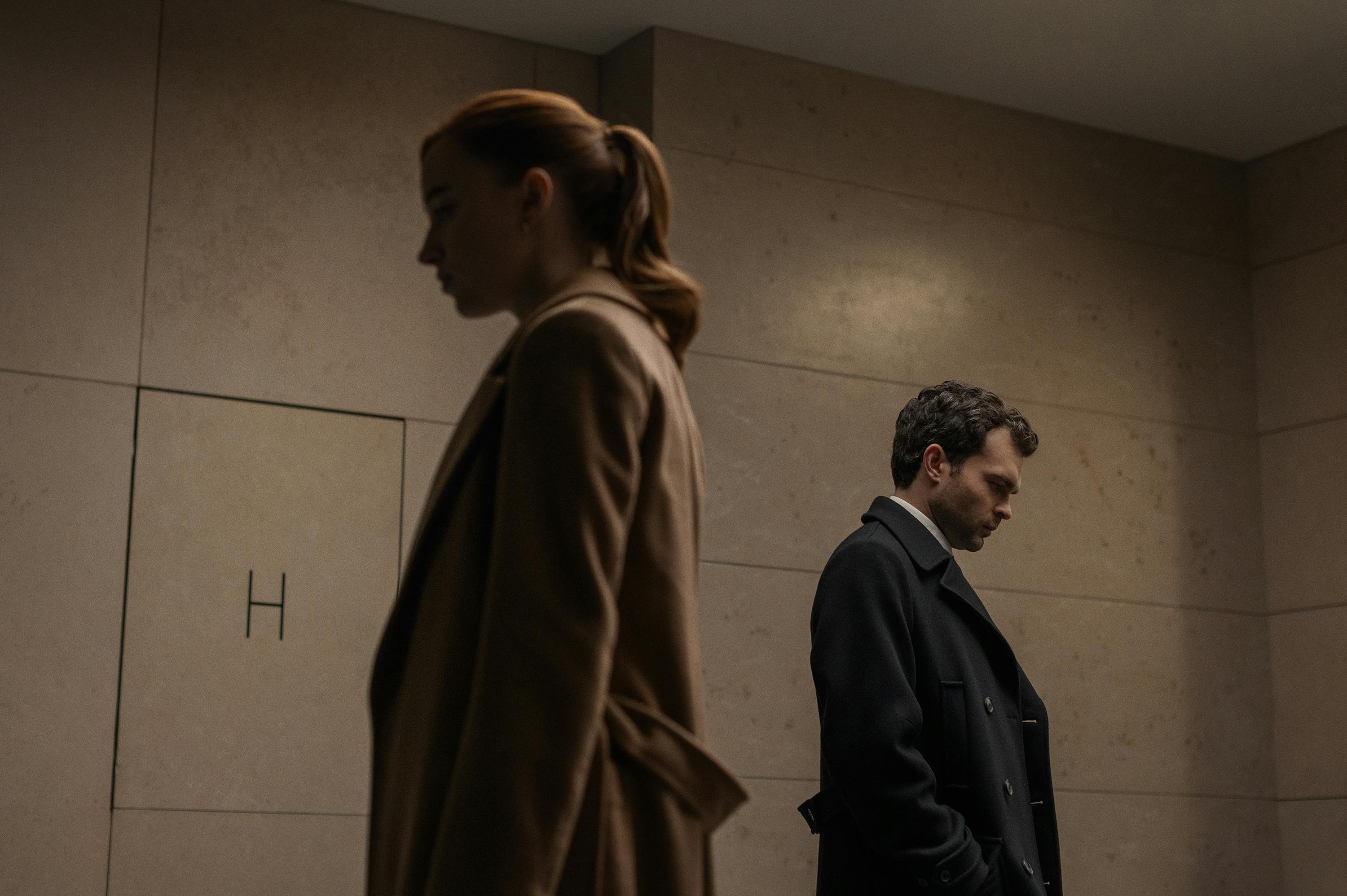 Emily (Phoebe Dynevor) and Luke (Alden Ehrenreich) pass by each other in this dimly-lit shot. Both actors' faces are in profile.