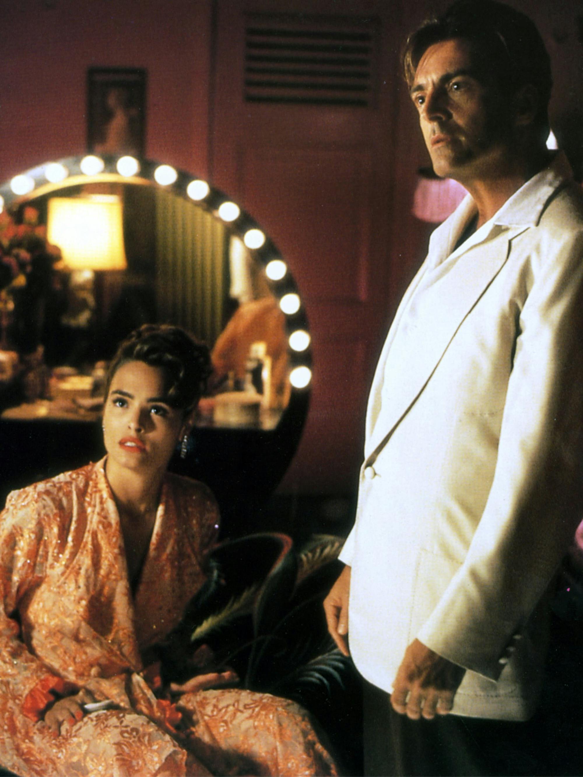 Talisa Soto sits in a robe. Behind her there is a circle-mirrored vanity with lights bordering the mirror. Armand Assante stands to the left. 