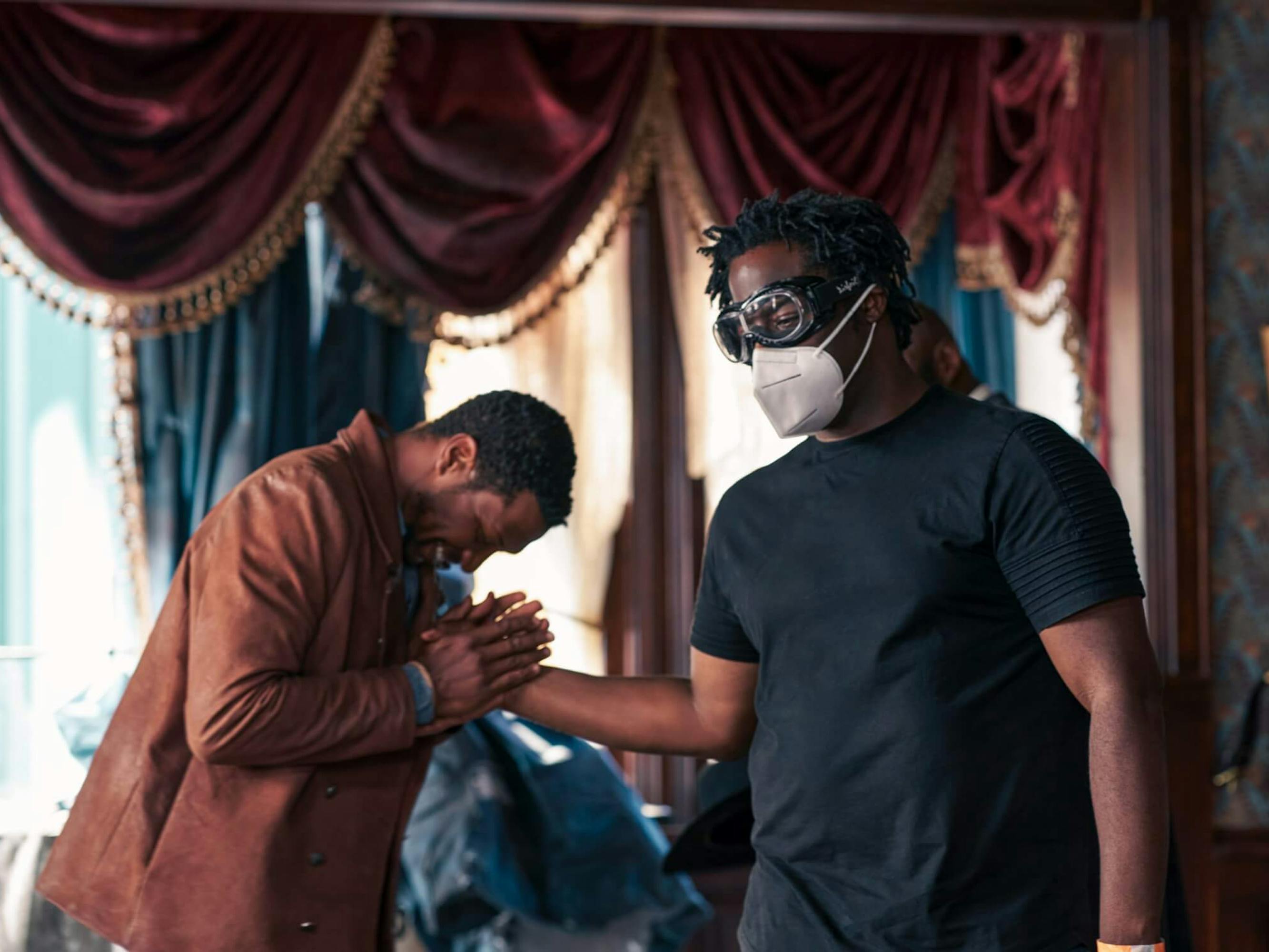 Jonathan Majors and Jeymes Samuel stand together in an ornately decorated room. Samuel wears all black, goggles, and a white mask. Majors holds Samuel hand in his and bends over it, as if in prayer. Majors wears a brown leather jacket.