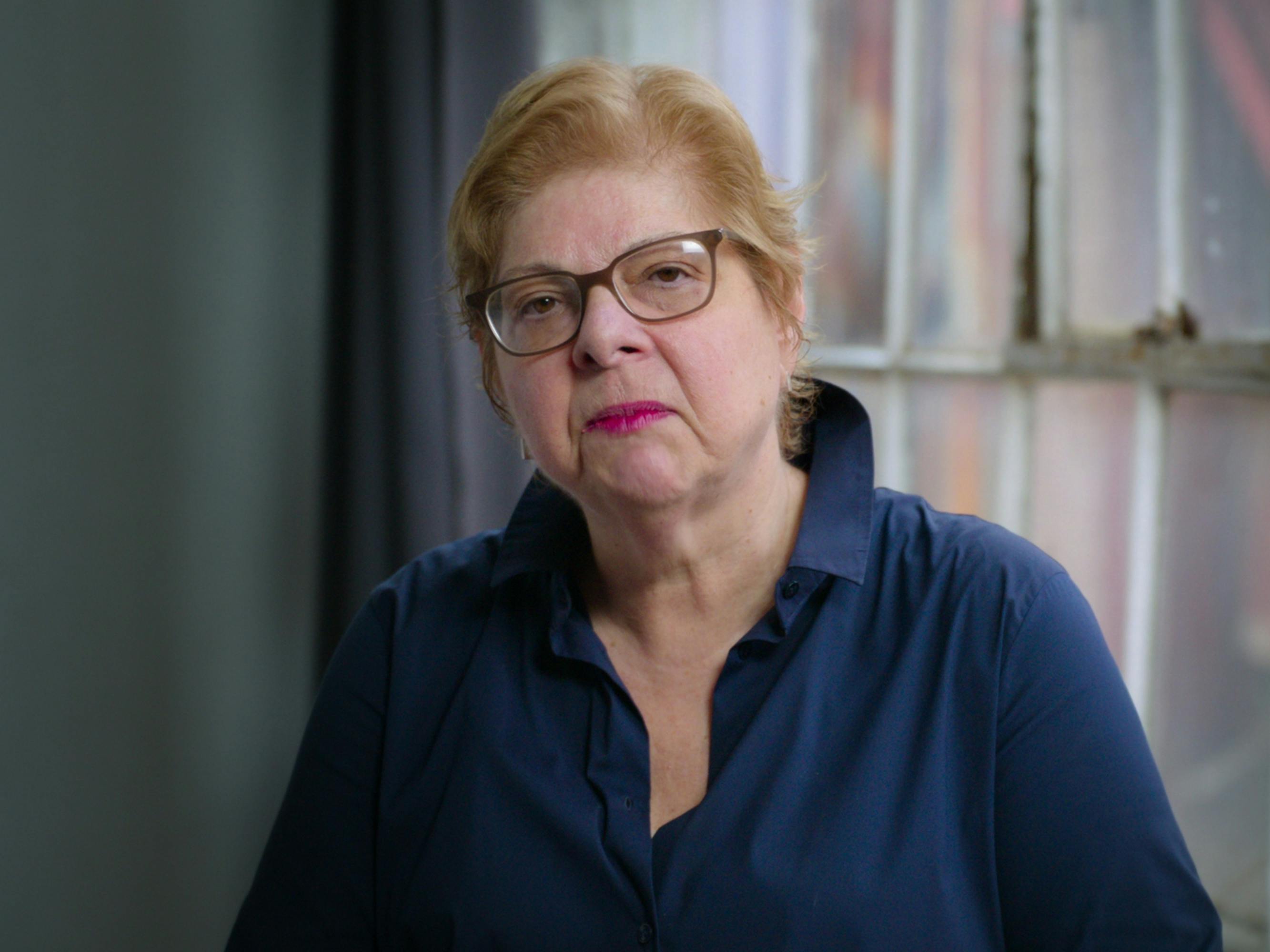 Donna de Salvo sits near a window and wears a navy shirt, glasses, and a pink lip.