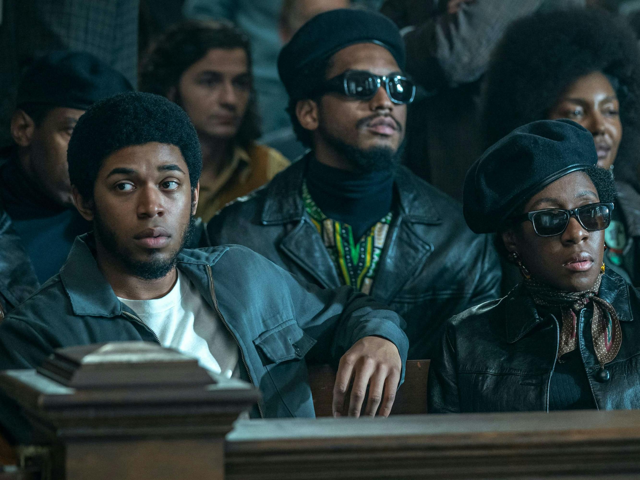 Harrison plays Black Panther Fred Hampton in The Trial of the Chicago 7. He wears a black jacket and white t-shirt, and he rests his elbow against a courtroom bench. He is surrounded by other people also wearing black.