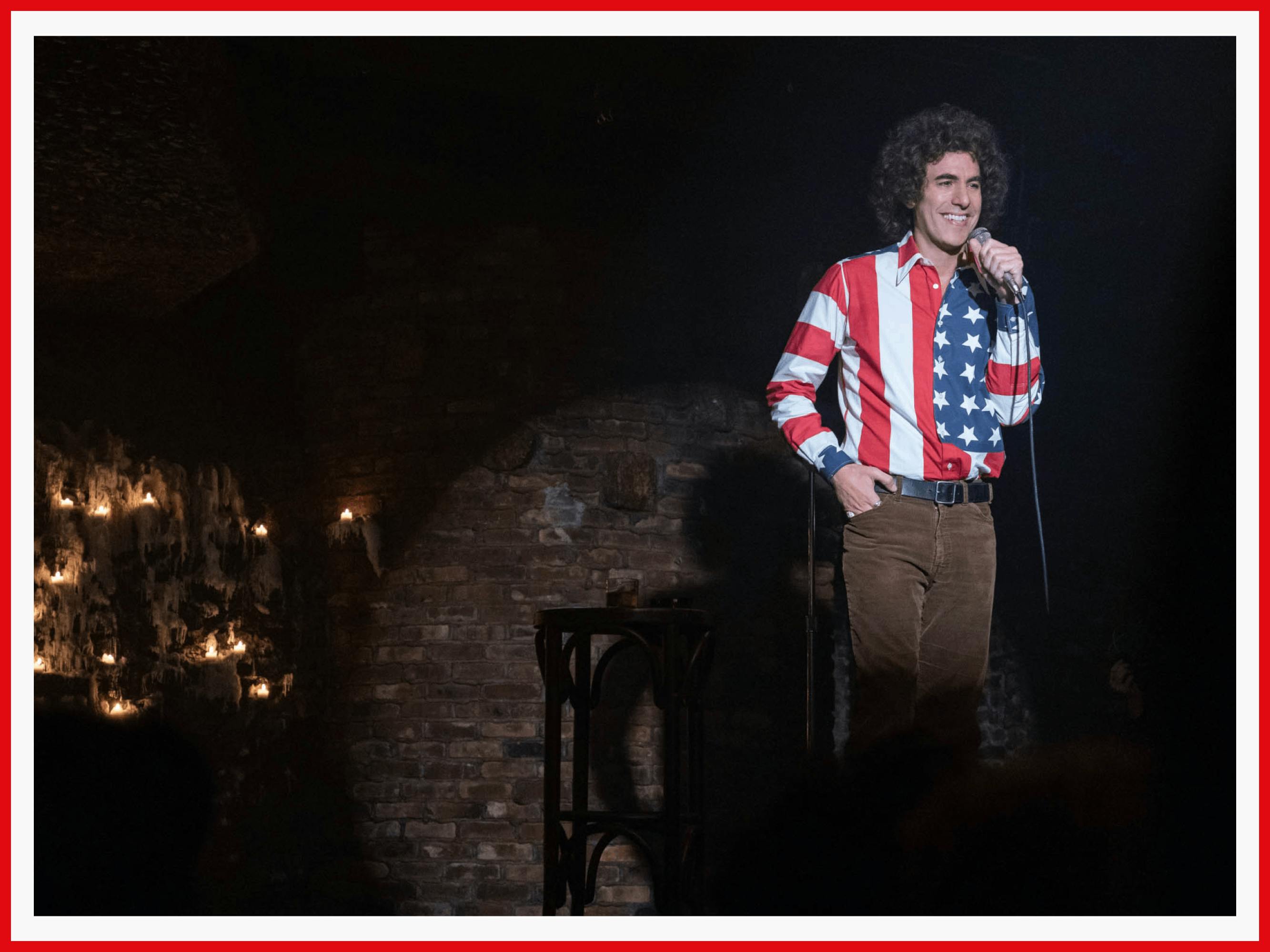 Sacha Baron Cohen, as Abbie Hoffman, stands grinning at a mic, wearing an extremely loud American-flag shirt. The sleeves are stripes, the cuffs are stars. 