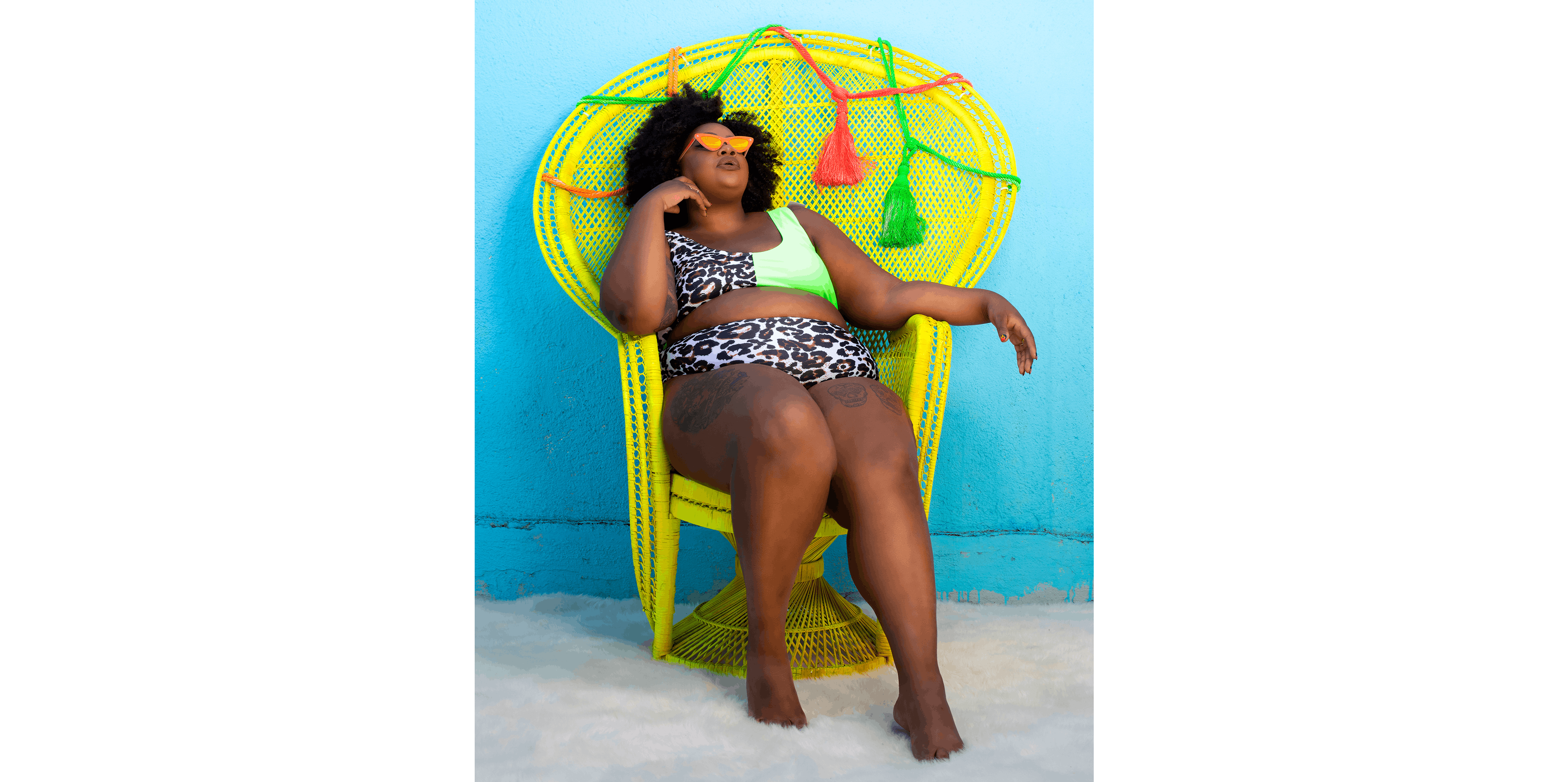 Nicole Byer basks in a bright yellow wicker chair in front of a sky blue wall, wearing a bikini with leopard print and sunglasses.