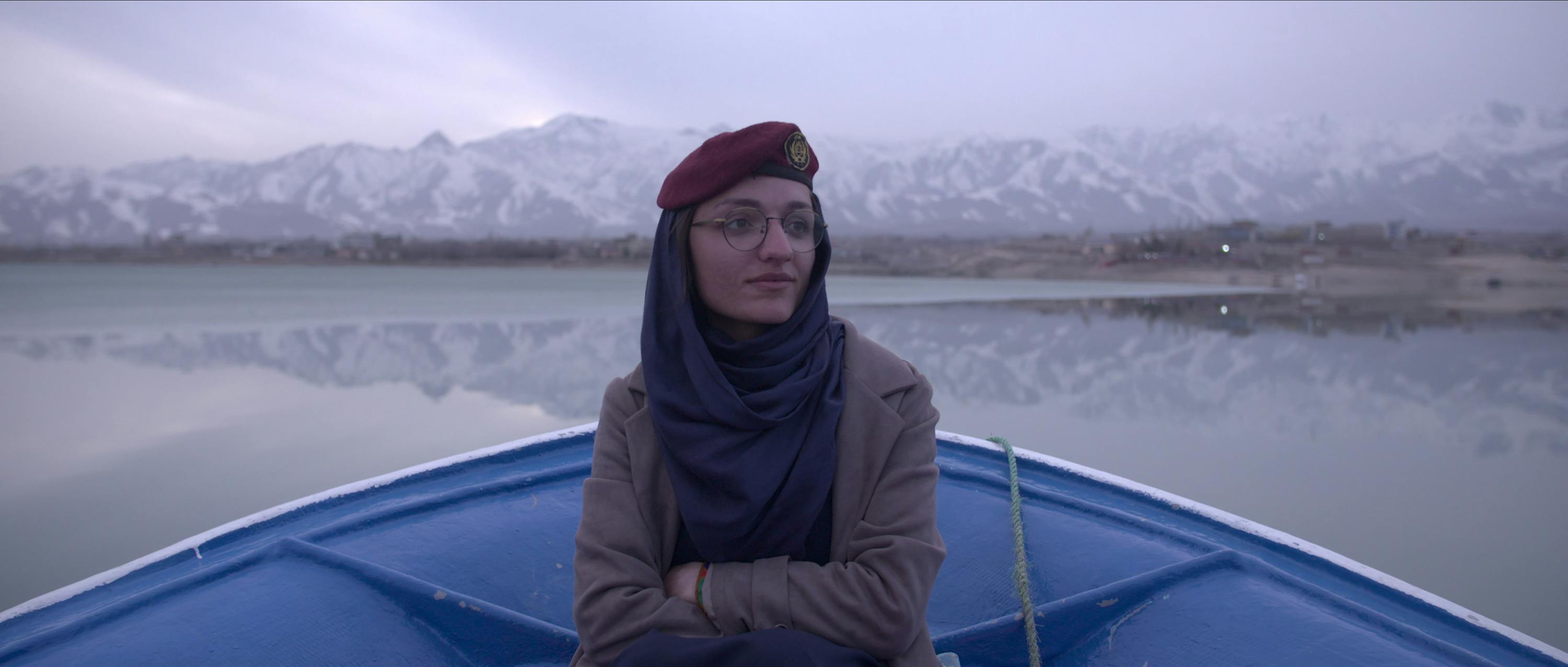 Zarifa Ghafari wears a grey jacket and sits in a blue boat, drifting on a very still body of water, framed by some mountains. 