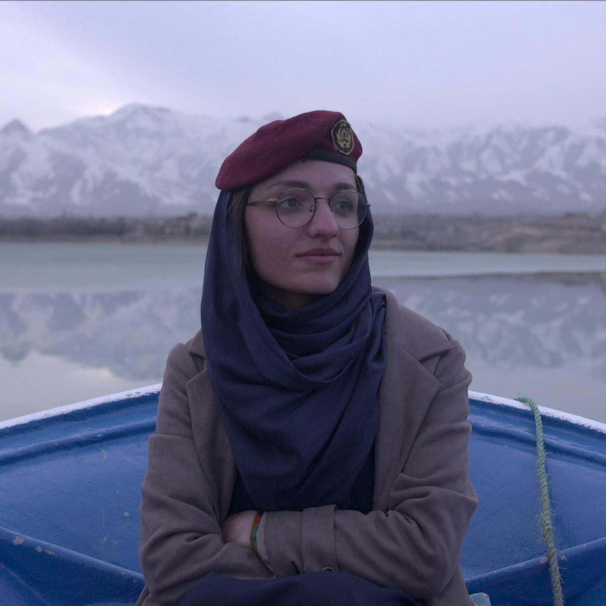 Zarifa Ghafari wears a grey jacket and sits in a blue boat, drifting on a very still body of water, framed by some mountains. 