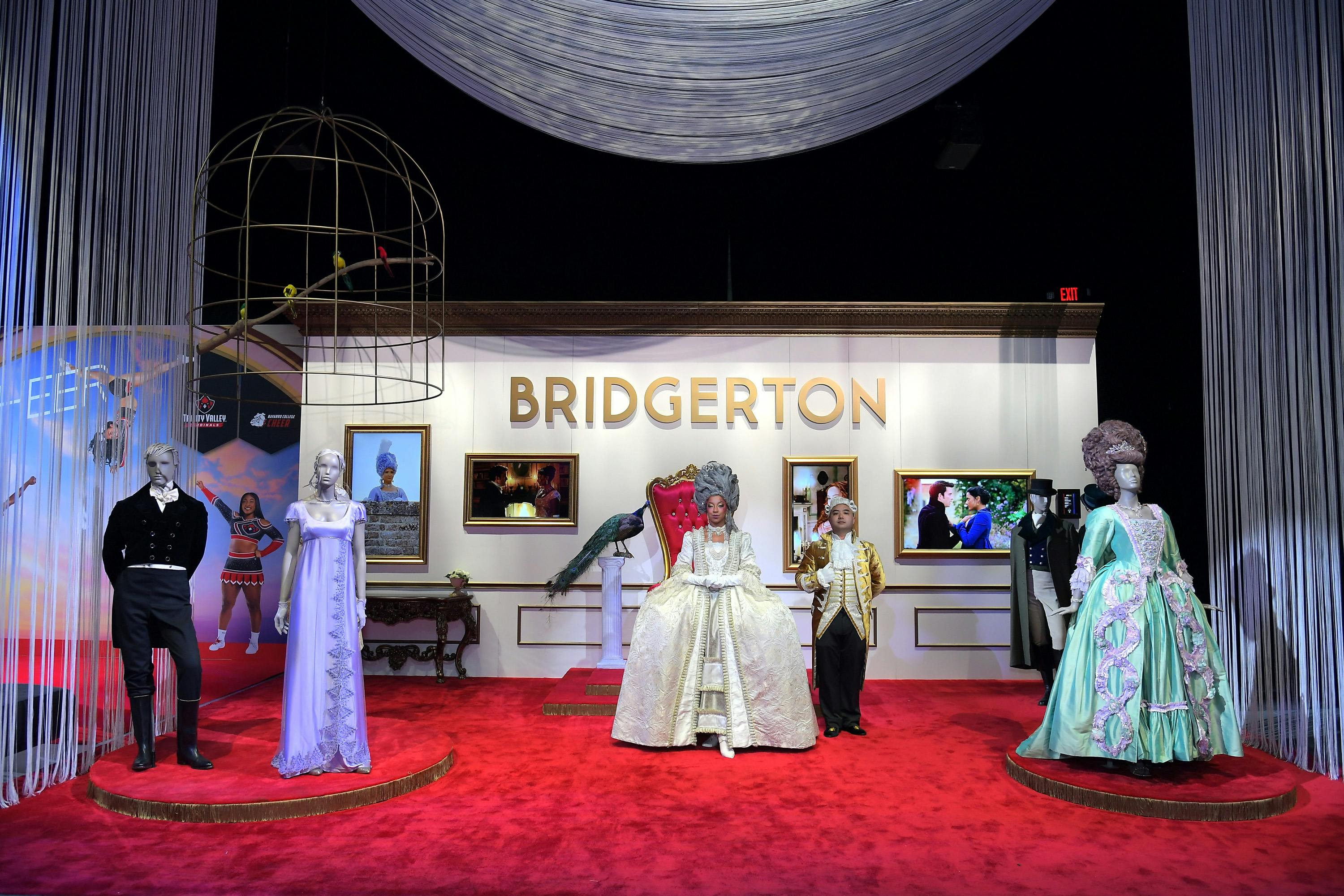 Dresses from season 2 of Bridgerton stand on a red plush carpet, against a wall that reads Bridgerton in gold.