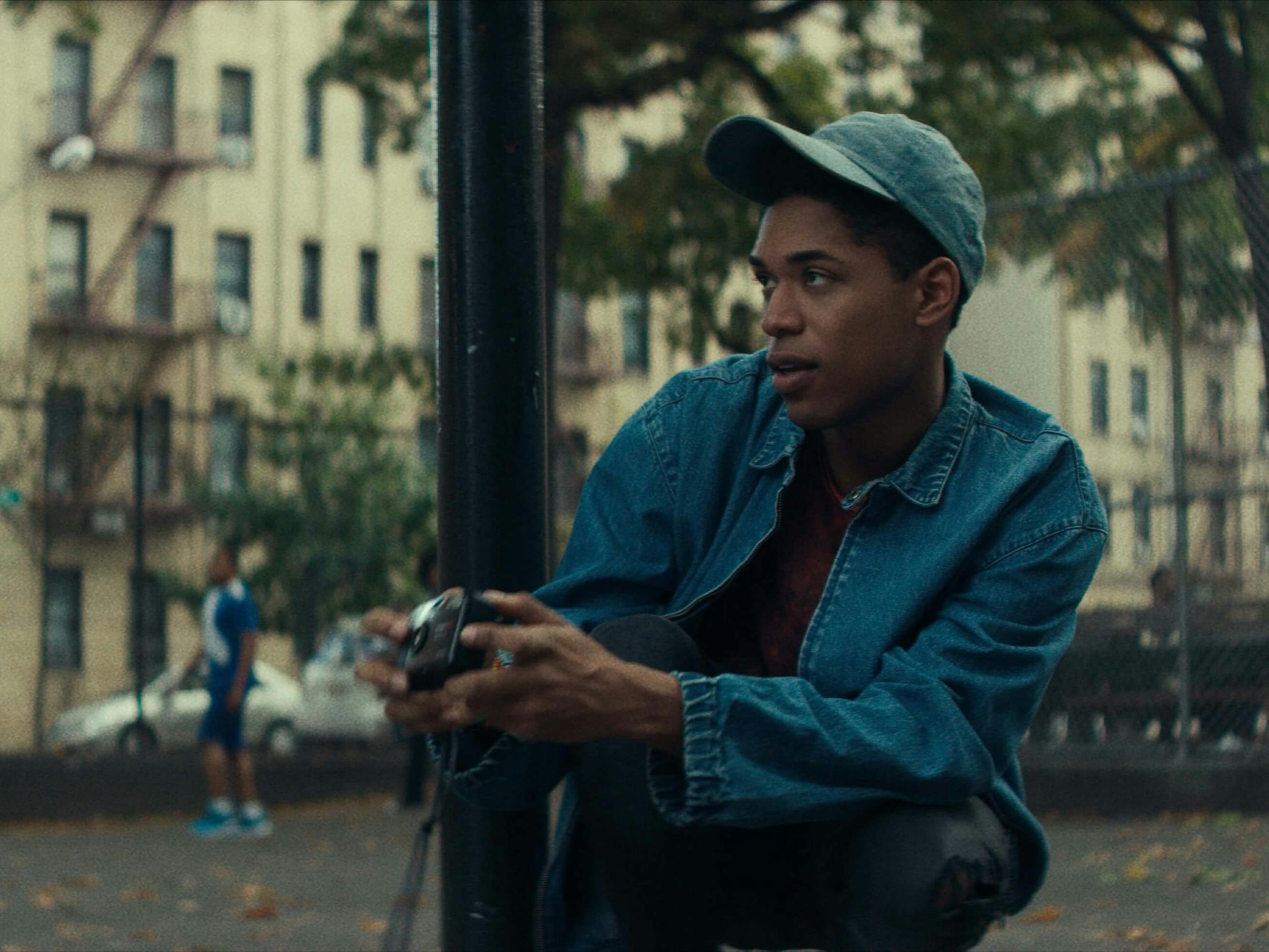 Harrison as Steven Harmon in Monster. He wears a jean jacket, grey hat, and he squats in what looks like a park. Apartment buildings loom behind him, and people play basketball in the background. 