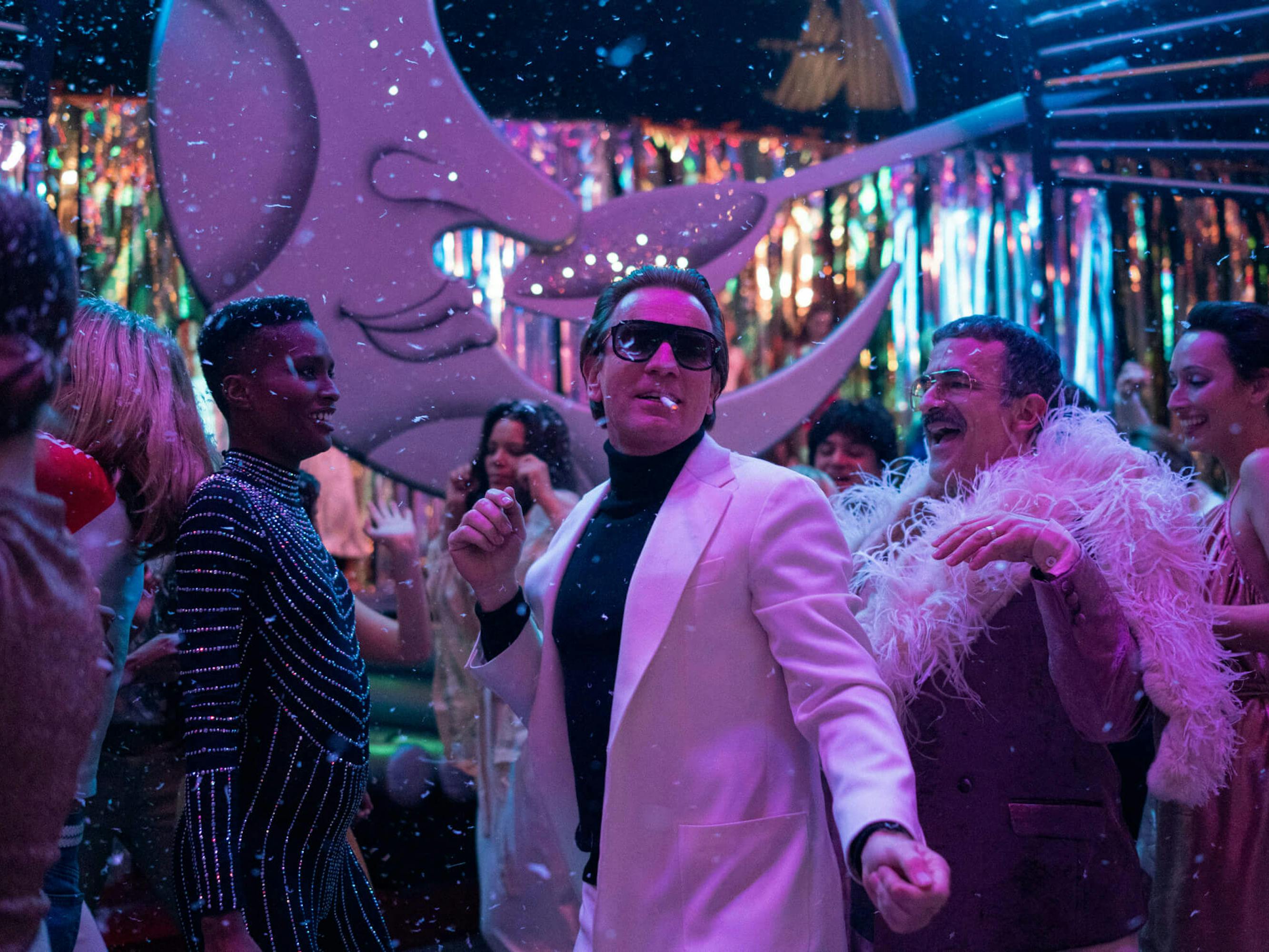 Halston (Ewan McGregor), Joe Eula (David Pittu), and Elsa Peretti (Rebecca Dayan) dance in Studio 54. Halston wears a white suit, Eula wears a feathered poncho, and Peretti wears a dress. The scene is lit by streamers and rainbow lights.