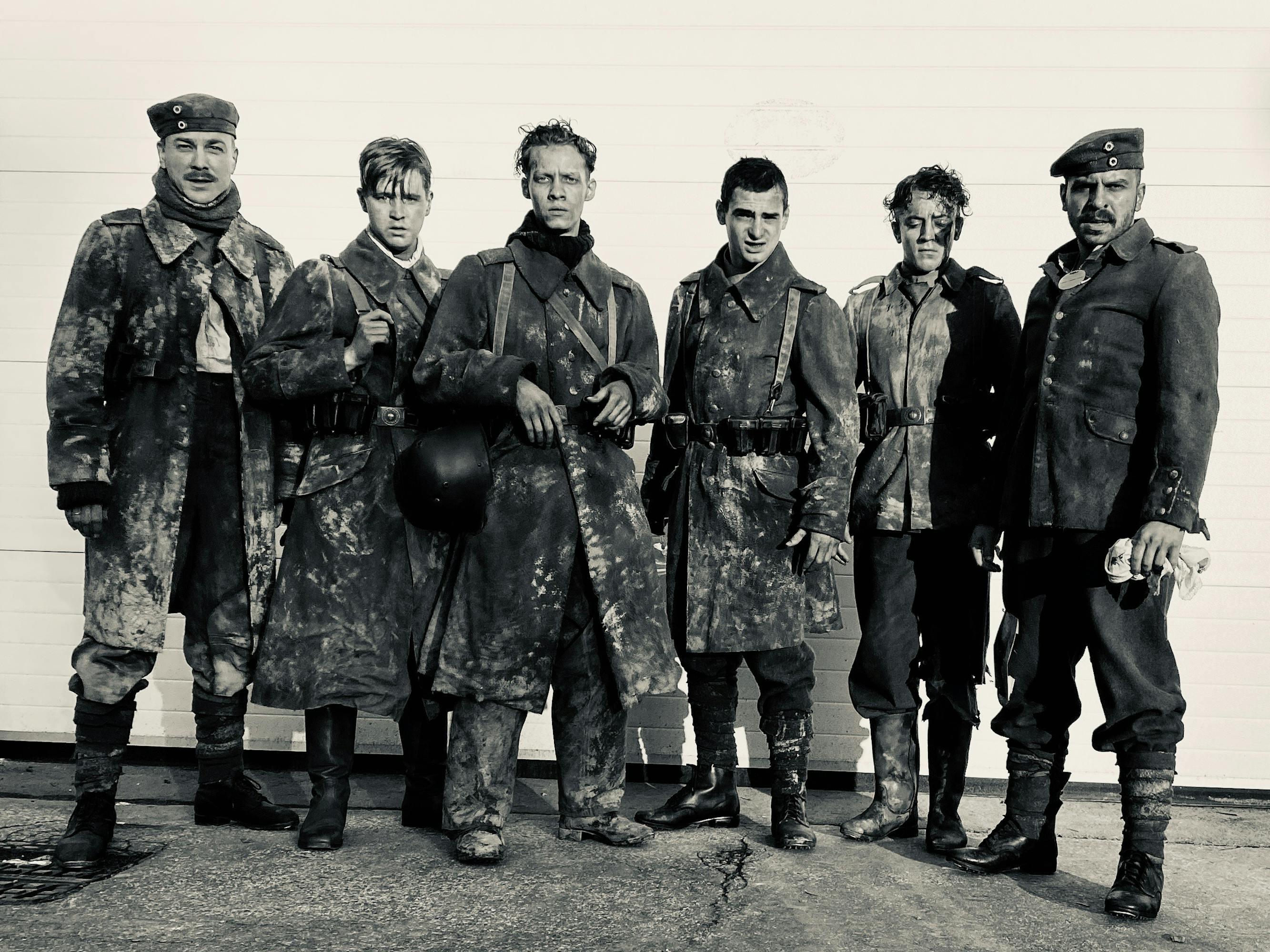 A line of soldiers stand together against a white wall. Their uniforms are muddied.