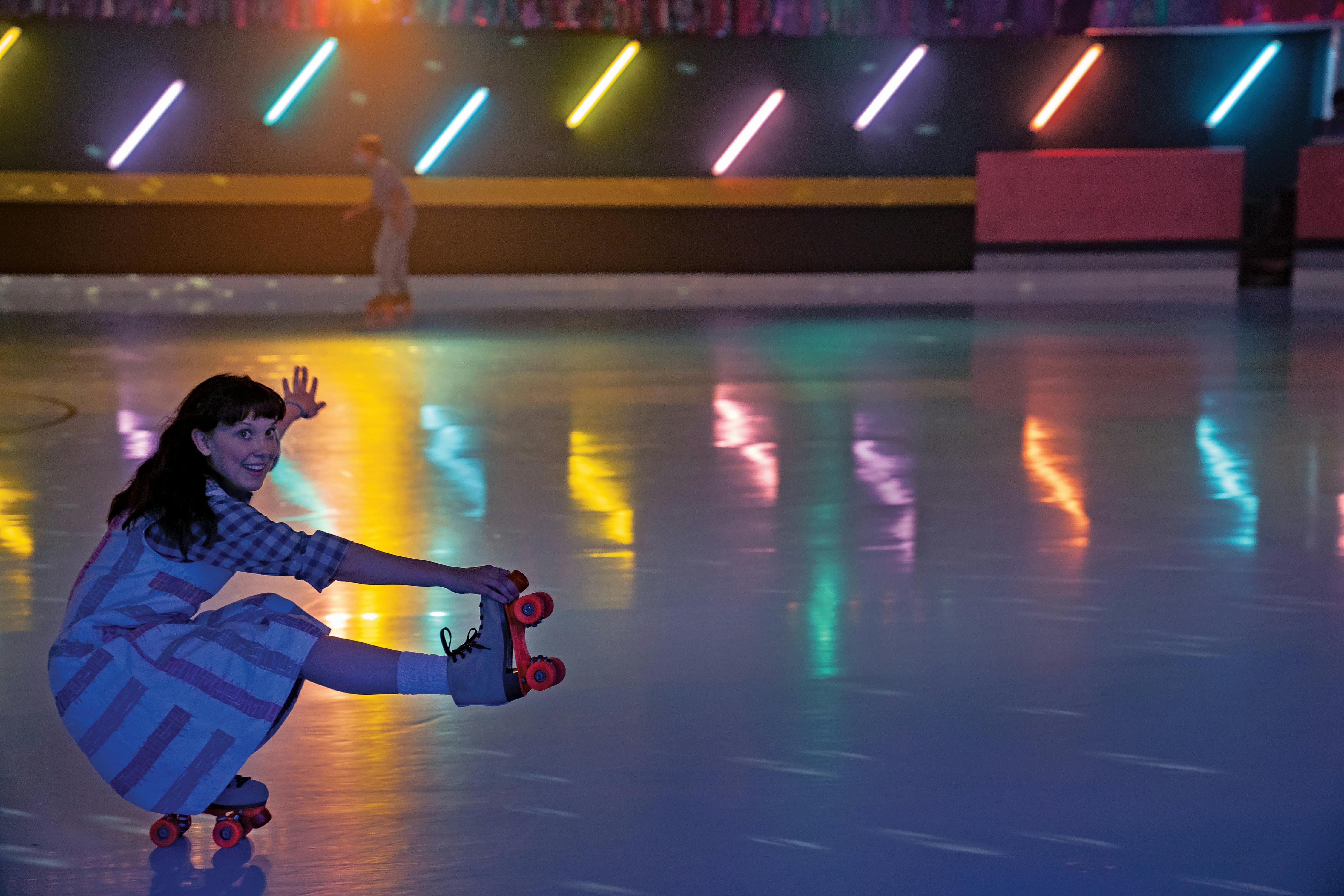 Millie Bobby Brown roller skates around a multi-colored rink in a patterned dress. 
