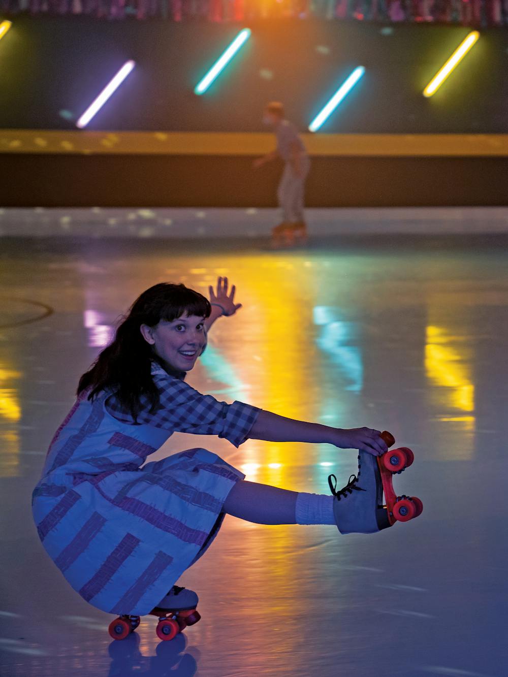 Millie Bobby Brown roller skates around a multi-colored rink in a patterned dress. 