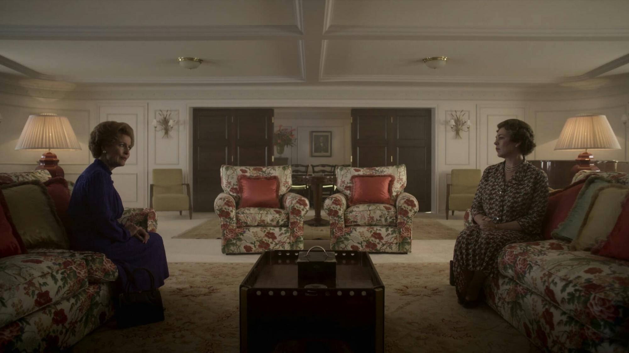 Margaret Thatcher (Gillian Anderson) and Queen Elizabeth (Olivia Colman) sit opposite each other on floral couches in a dimly lit room.