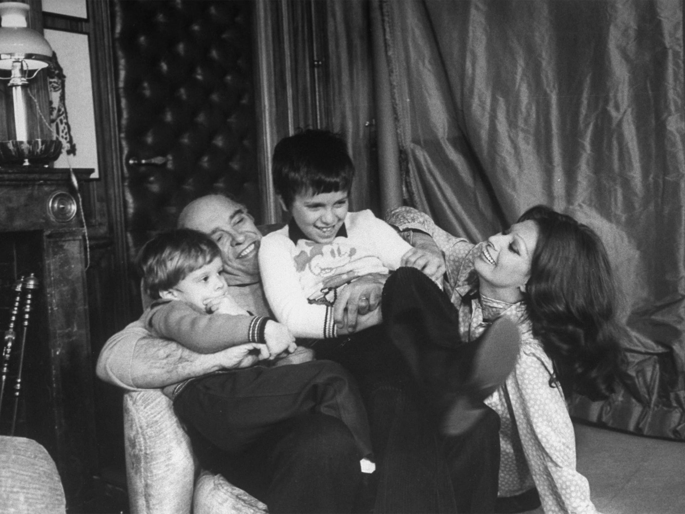 In this playful old photograph, Carlo Ponti sits in an armchair, holding his two young children, while Loren smiles at them from her seat on the floor. 