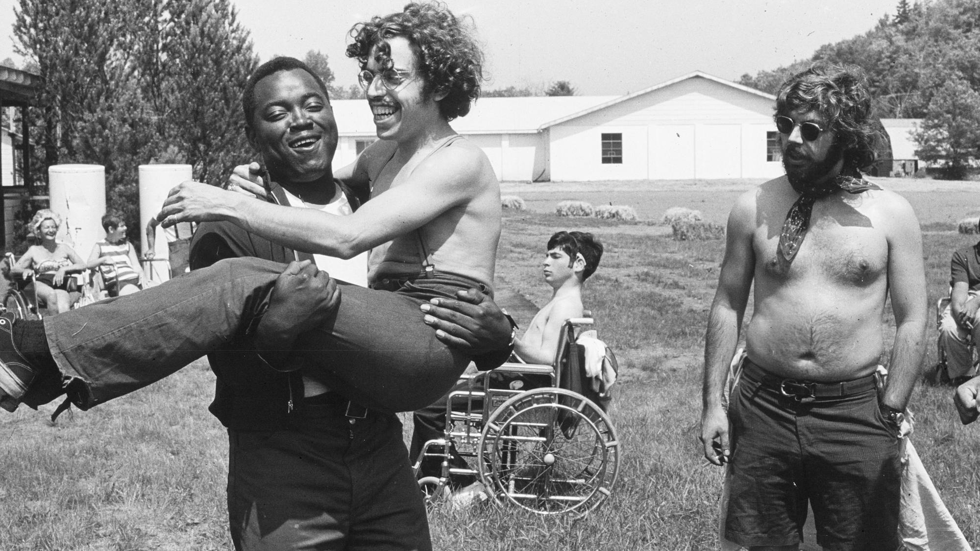 A black-and-white photograph of teenagers at camp. In the foreground, one camper carries his wheelchair-bound friend, both laughing, while another camper, shirtless and sporting a neckerchief, looks on.
