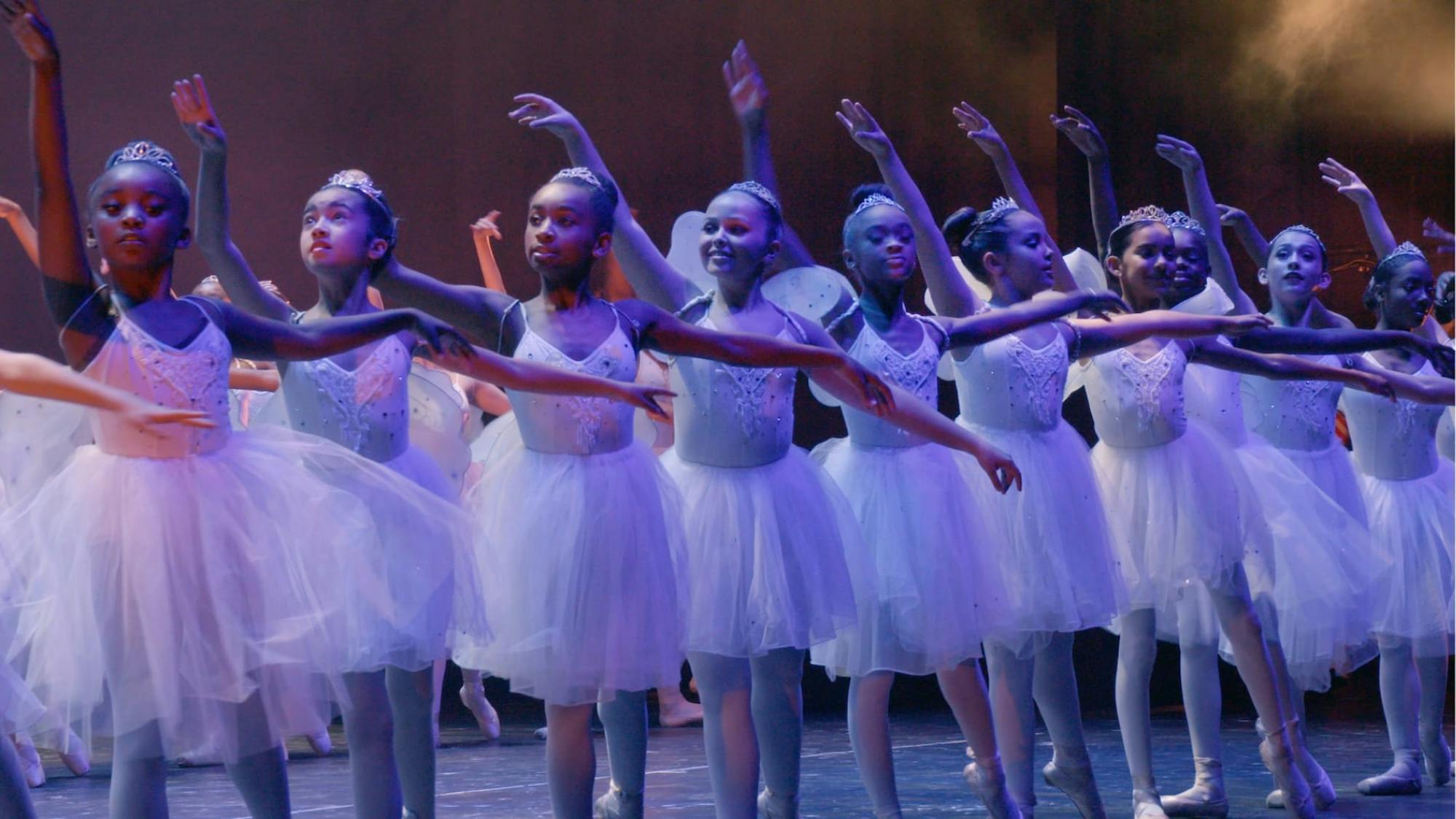 Young ballerinas stand in a line wearing tutus, lit by a blue light.