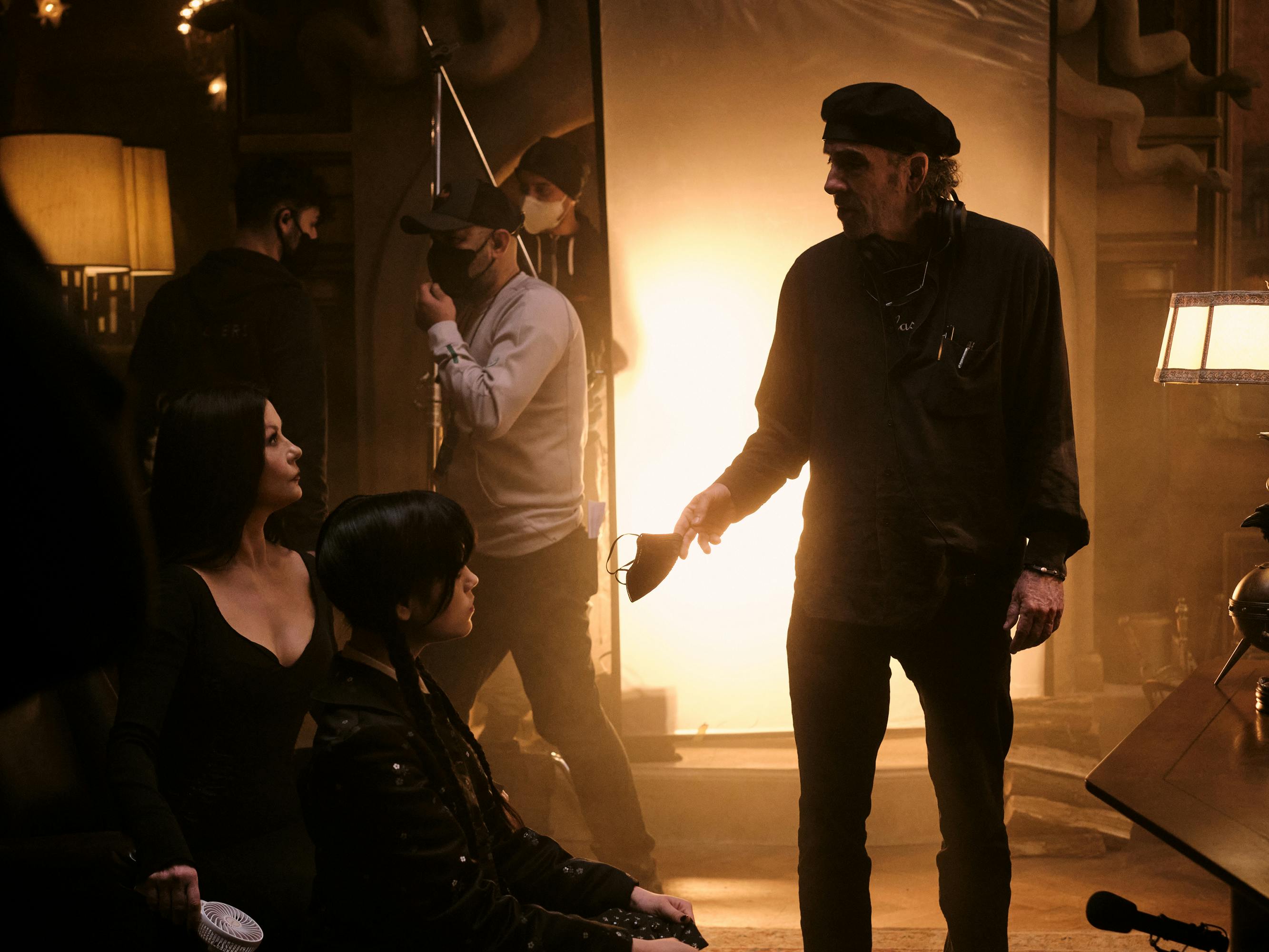 Catherine Zeta-Jones and Jenna Ortega sit across from Tim Burton as he stands, lit from behind by a bright golden light.