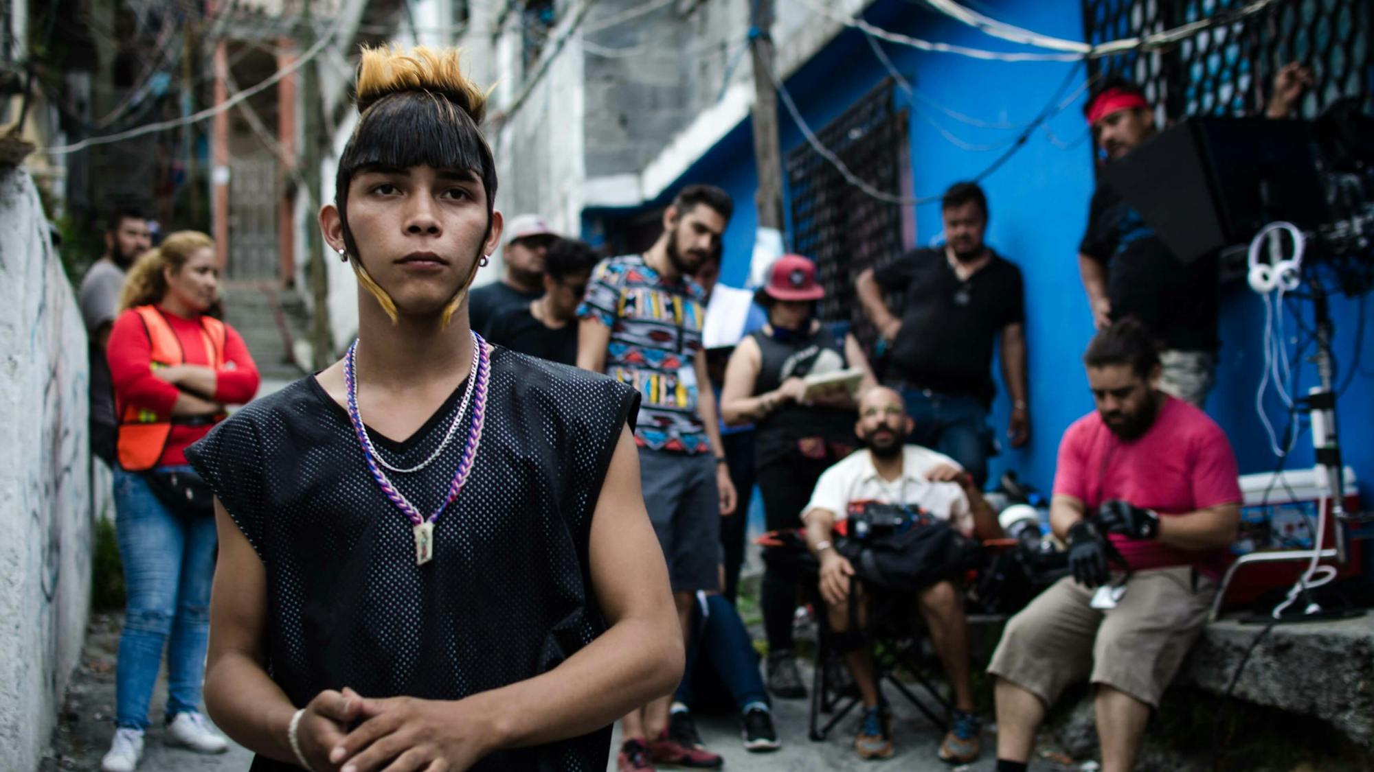 Juan Daniel Garcia Treviño in costume as Ulises, with his signature gelled hairstyle. He stands in a Monterrey alleyway with the film’s crew behind him.