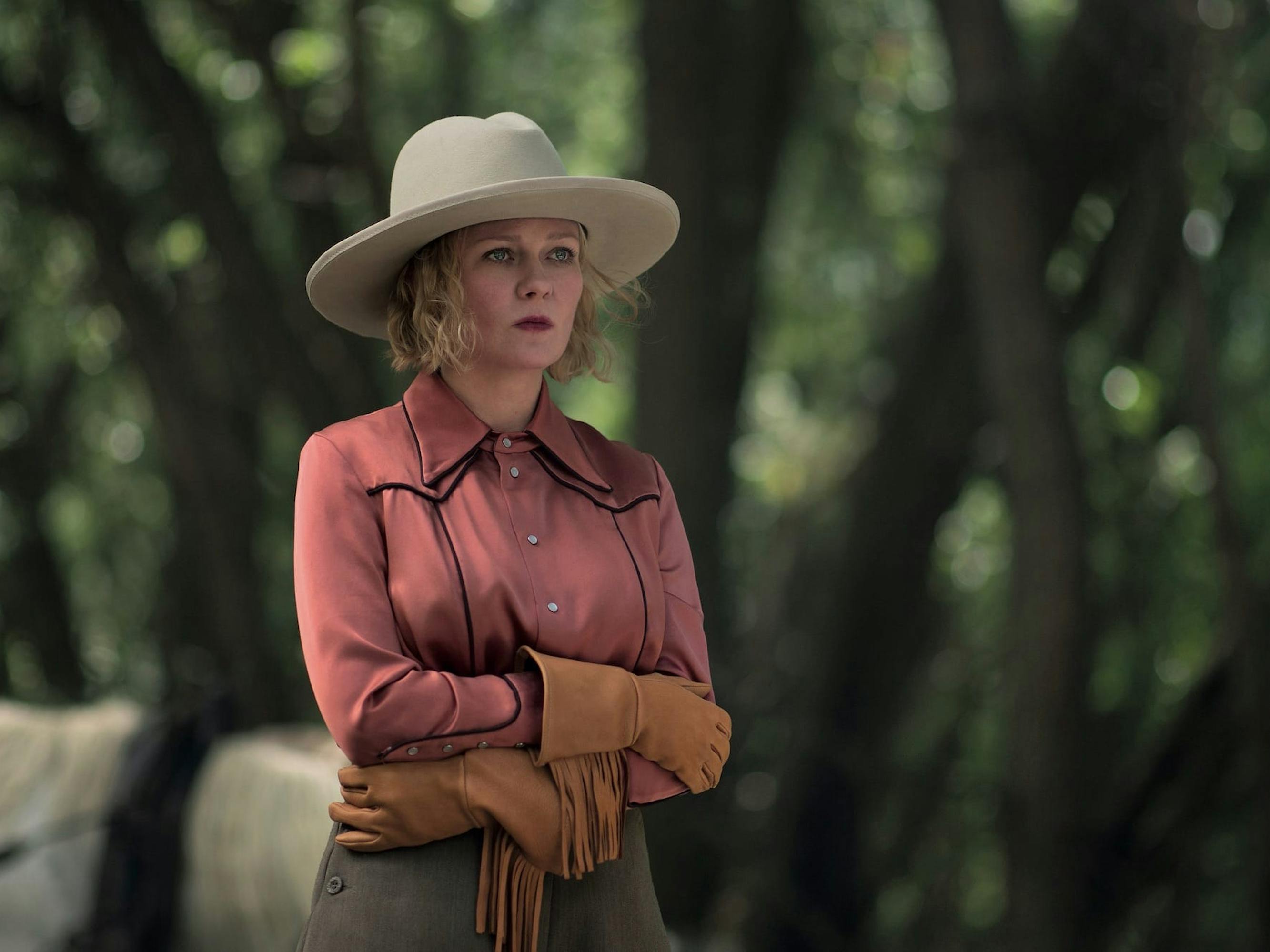 Rose (Kirsten Dunst) wears a red shirt, tan hat, and brown gloves. Behind her are dappled green trees.