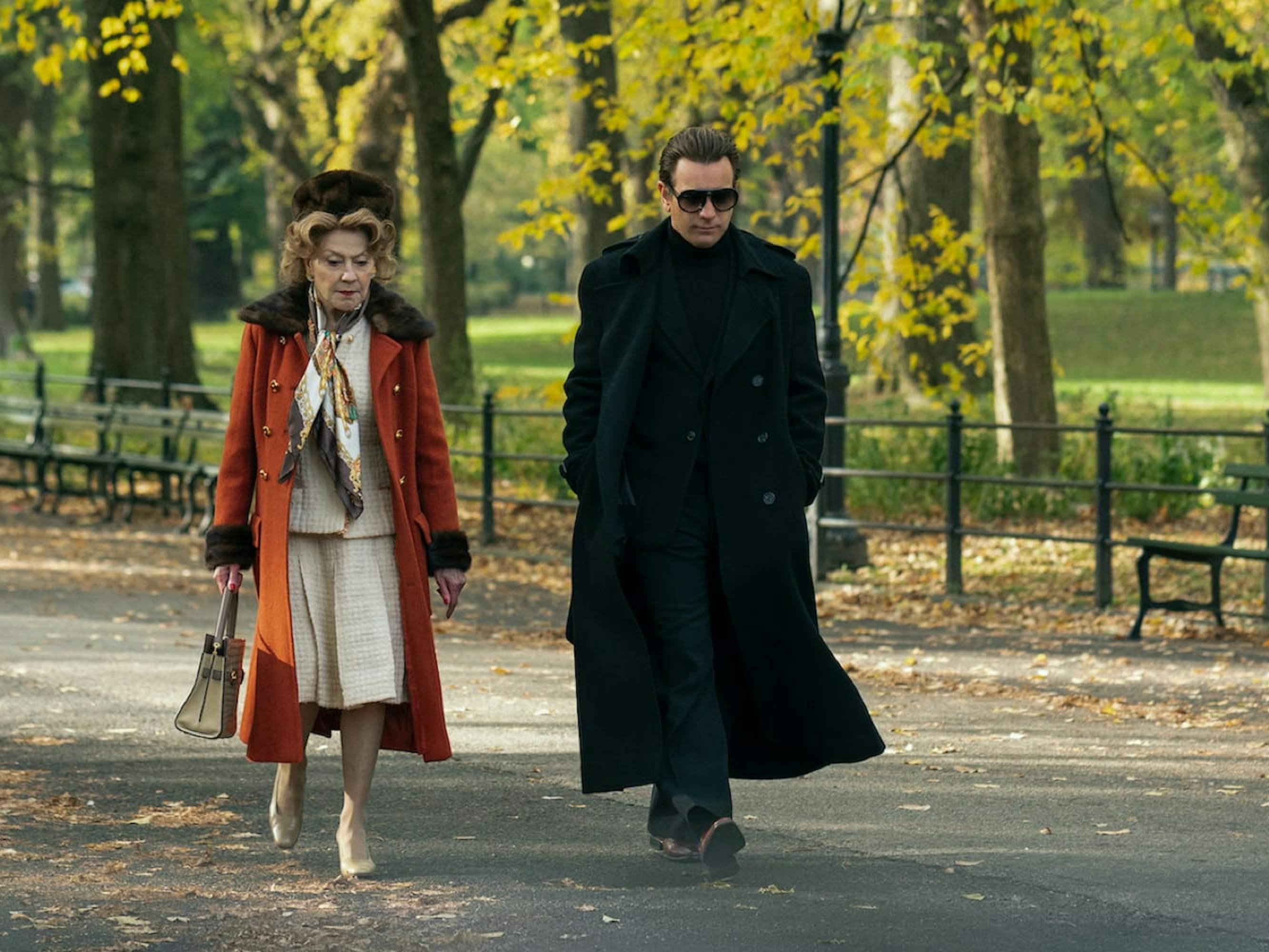 Eleanor Lambert (Kelly Bishop) and Halston (Ewan McGregor) walk in Central Park. Lambert wears a glorious orange coat trimmed with fur. Halston stands out in his signature all black look, including sunglasses. The fall leaves, trees, and empty benches are reminiscent of When Harry Met Sally’s epic shot.