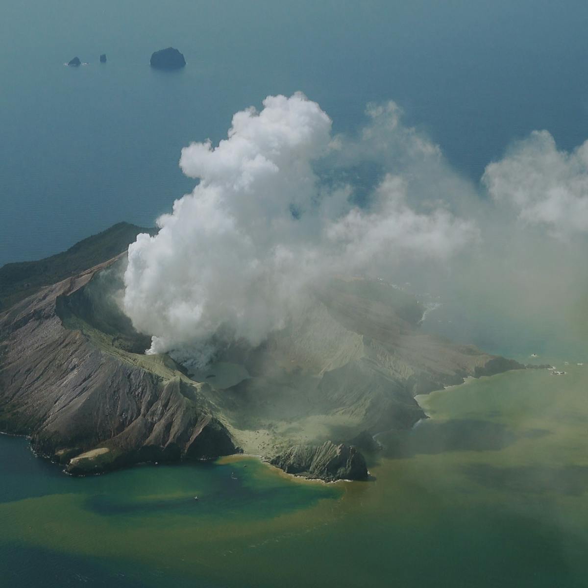 A smoking volcano amidst a yellow-blue-green ocean.