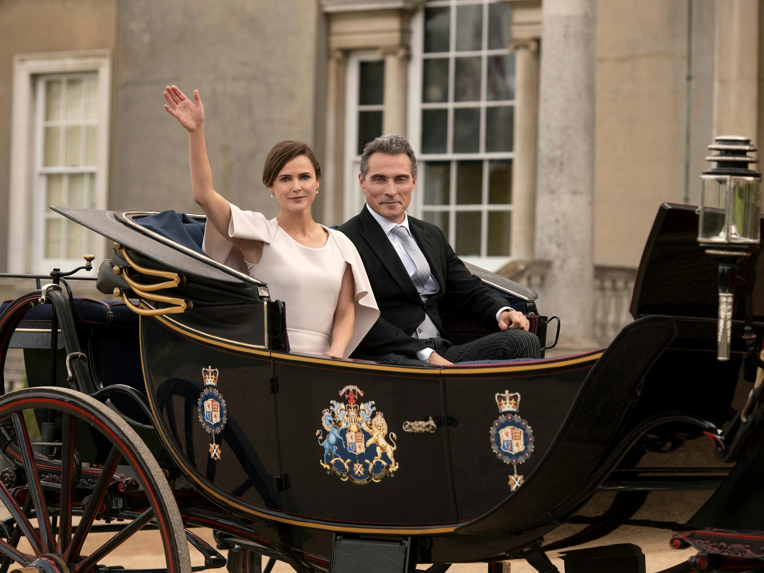 Kate Wyler (Keri Russell) and Hal Wyler (Rufus Sewell) sit in a royal carriage. Kate wears a white dress and Hal wears a suit.