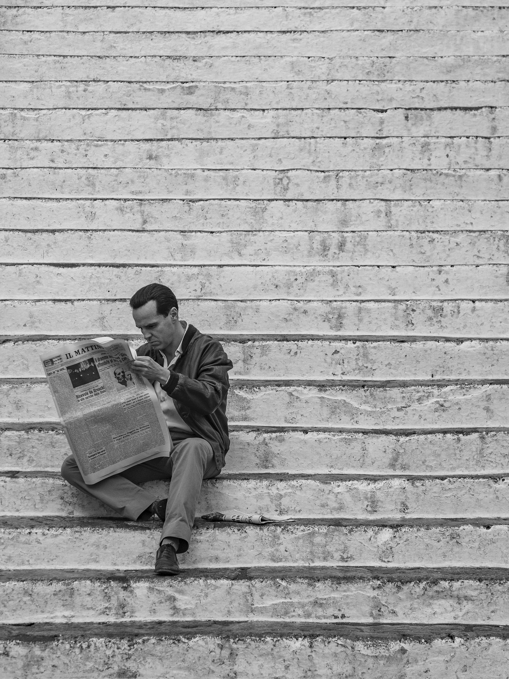Tom Ripley (Andrew Scott) sits on some stairs and reads a newspaper.