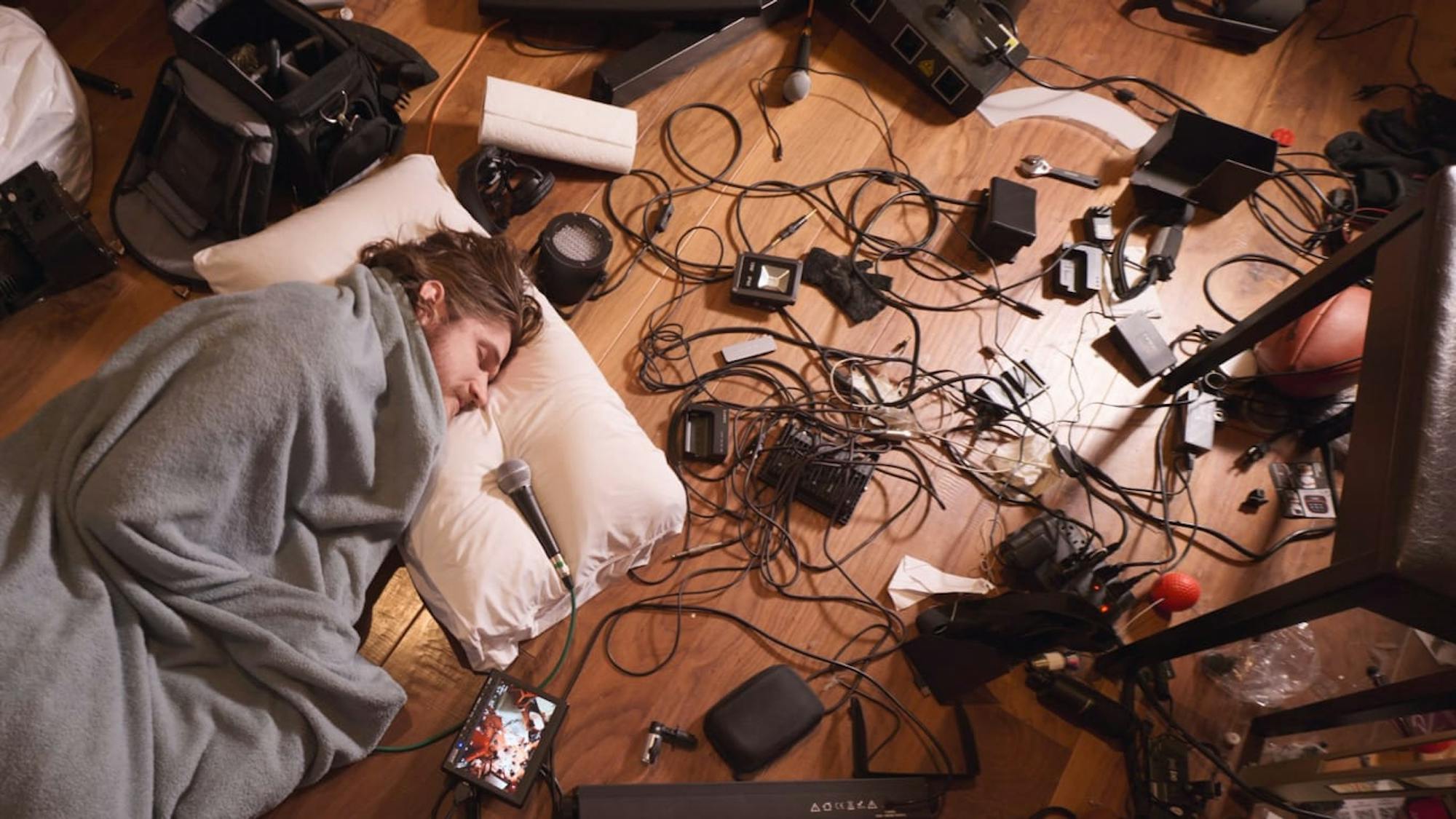 Bo Burnham sleeps on his hardwood floor, head resting against his white pillow. A mike lies pointing towards his mouth, and the floor is covered with cords and machines. Bo is wrapped in a grey blanket. The mess is chaotic, including a basketball, ipad, and stool.
