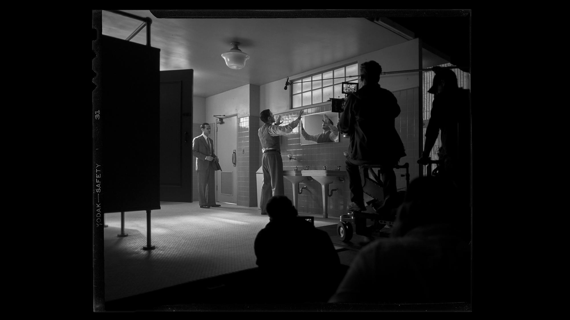 This behind-the-scenes shot shows the set of the men’s room at M-G-M. Tiles stretch up three quarters of one wall, and catch in the light. The crew is visible just off set.