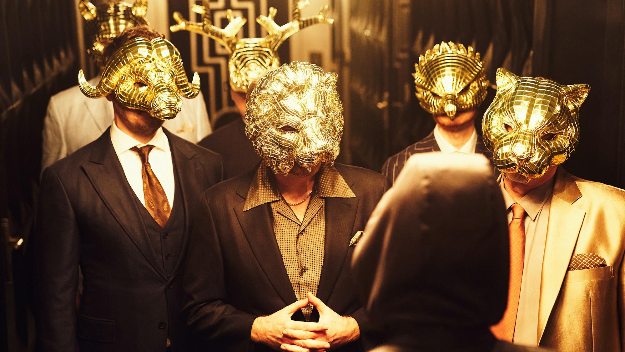 A group of V.I.P.s wear creepy gold animal masks and suits.