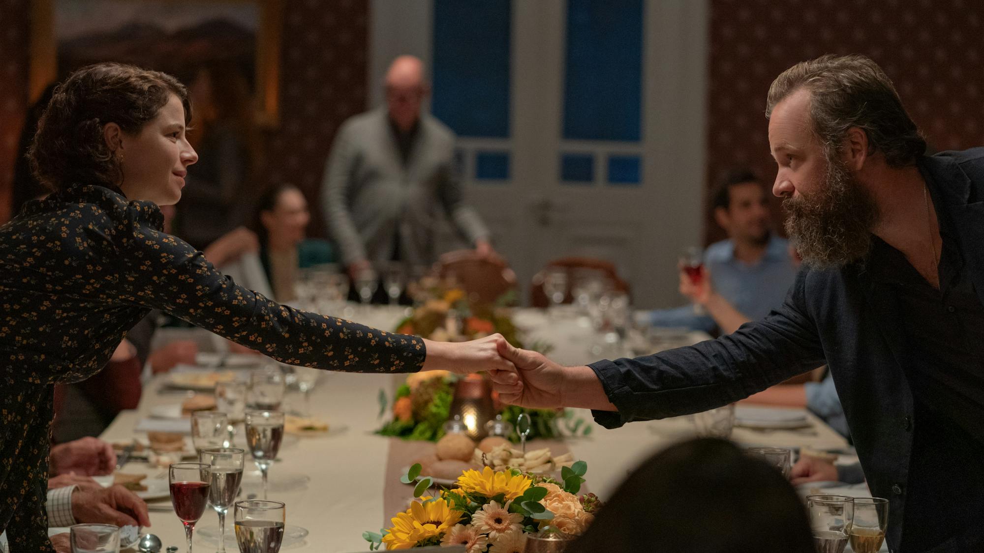 Jessie Buckley and Peter Sarsgaard shake hands over a table filled with cups, food, and flowers.