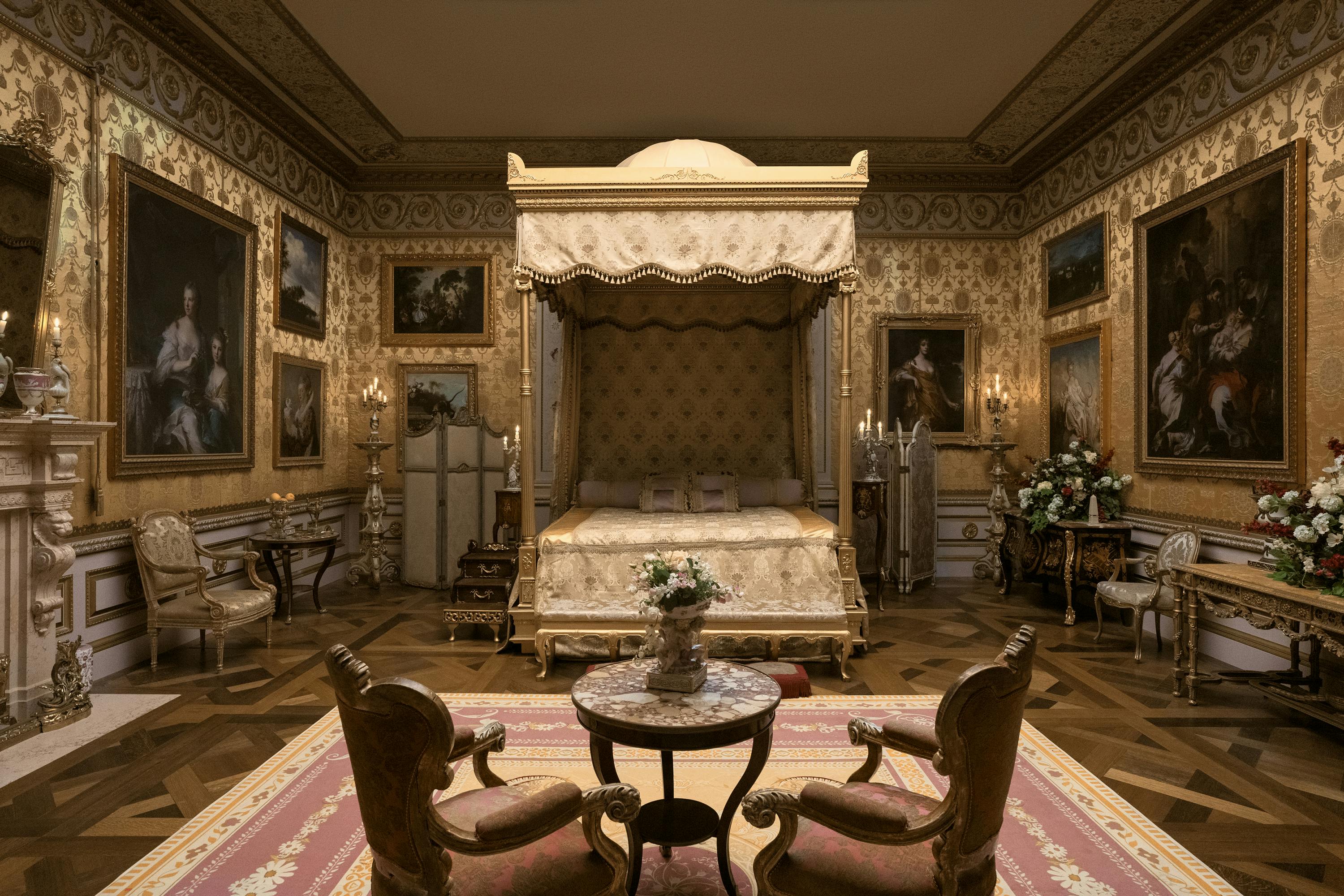 The monarch’s bedroom. A four-poster bed sits in the middle of the room. There is also a small table with two armchairs, and the walls are covered with ornate wallpaper and paintings. The color scheme is red, gold, and brown, and while the room looks cozy, its also very ornate and formal.