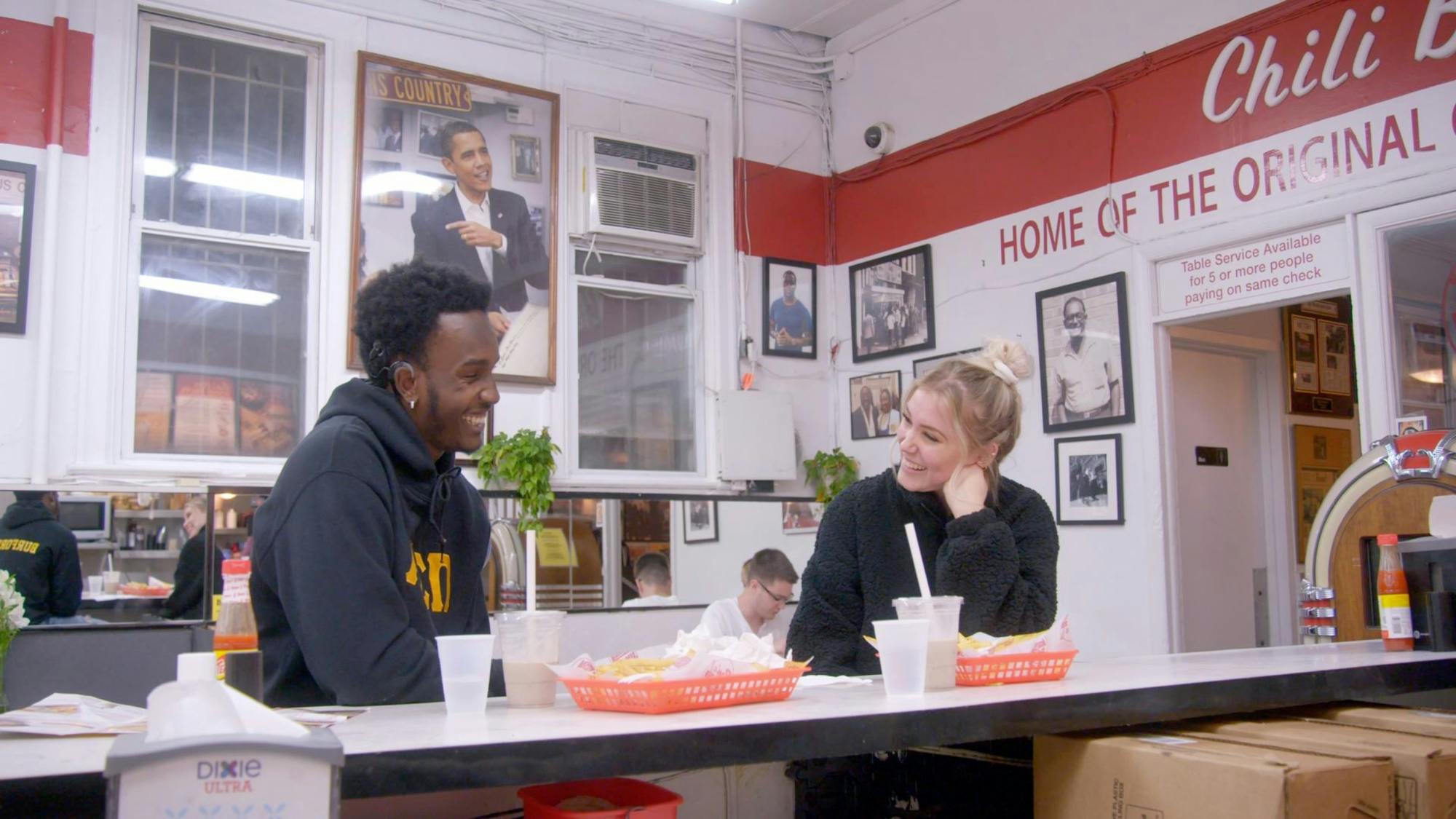 Cheyenna Clearbrook and Rodney Burford sit it in an old timey diner together. They both eat shakes and fries and are dressed in black sweatshirts. They smile at each other and look utterly adorable. In the background is a picture of President Barak Obama.