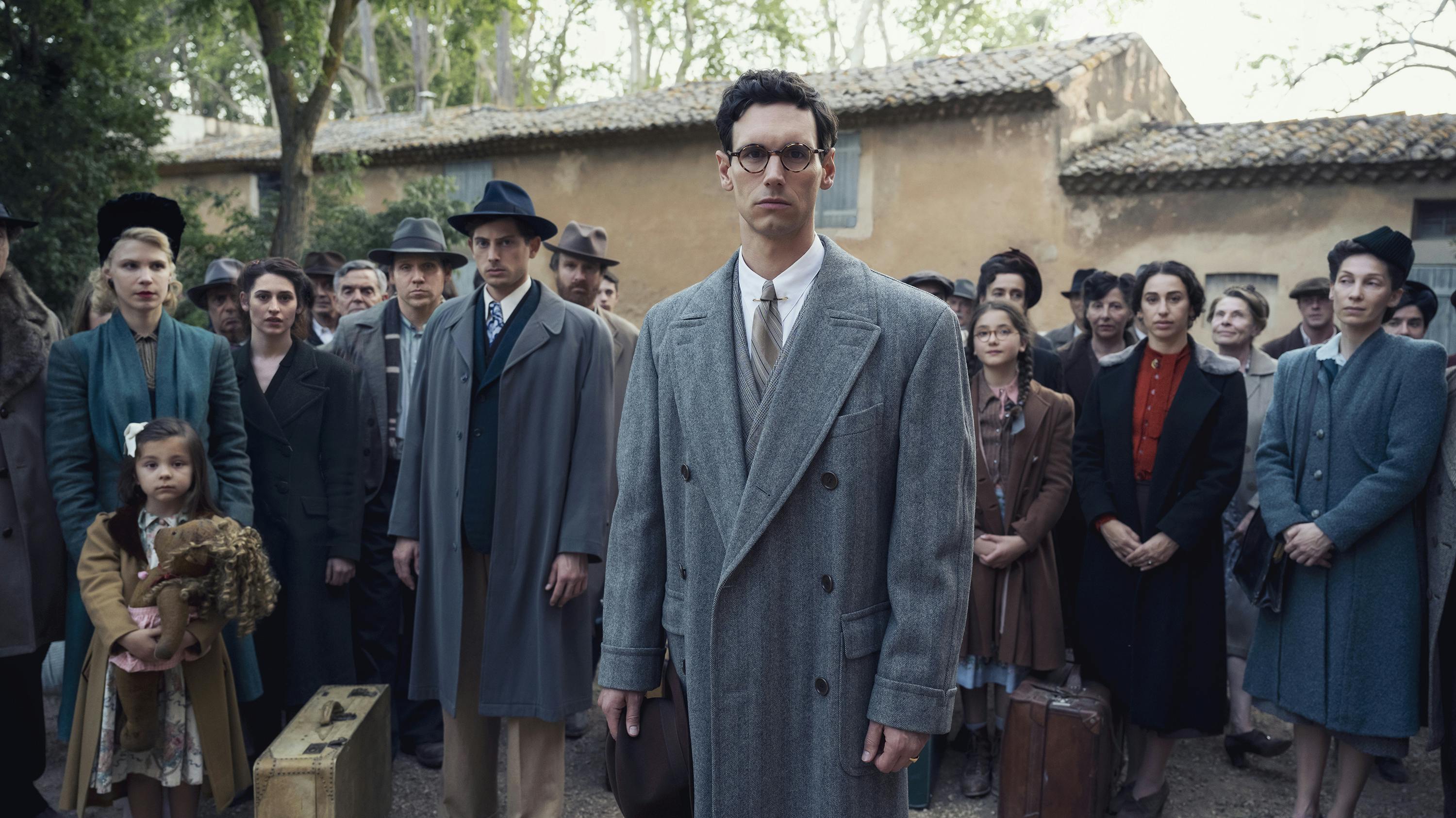Varian Fry (Cory Michael Smith) and Thomas Lovegrove (Amit Rahav) and other cast members stand in a line facing the camera wearing a range of blue, grey, and black coats.