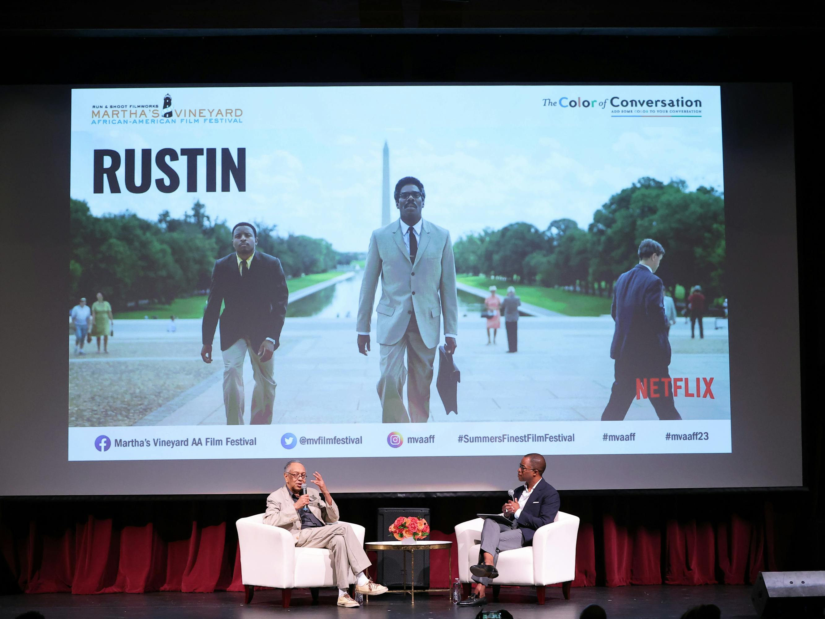 Two people sit onstage discussing Rustin, the title of which is projected behind them.