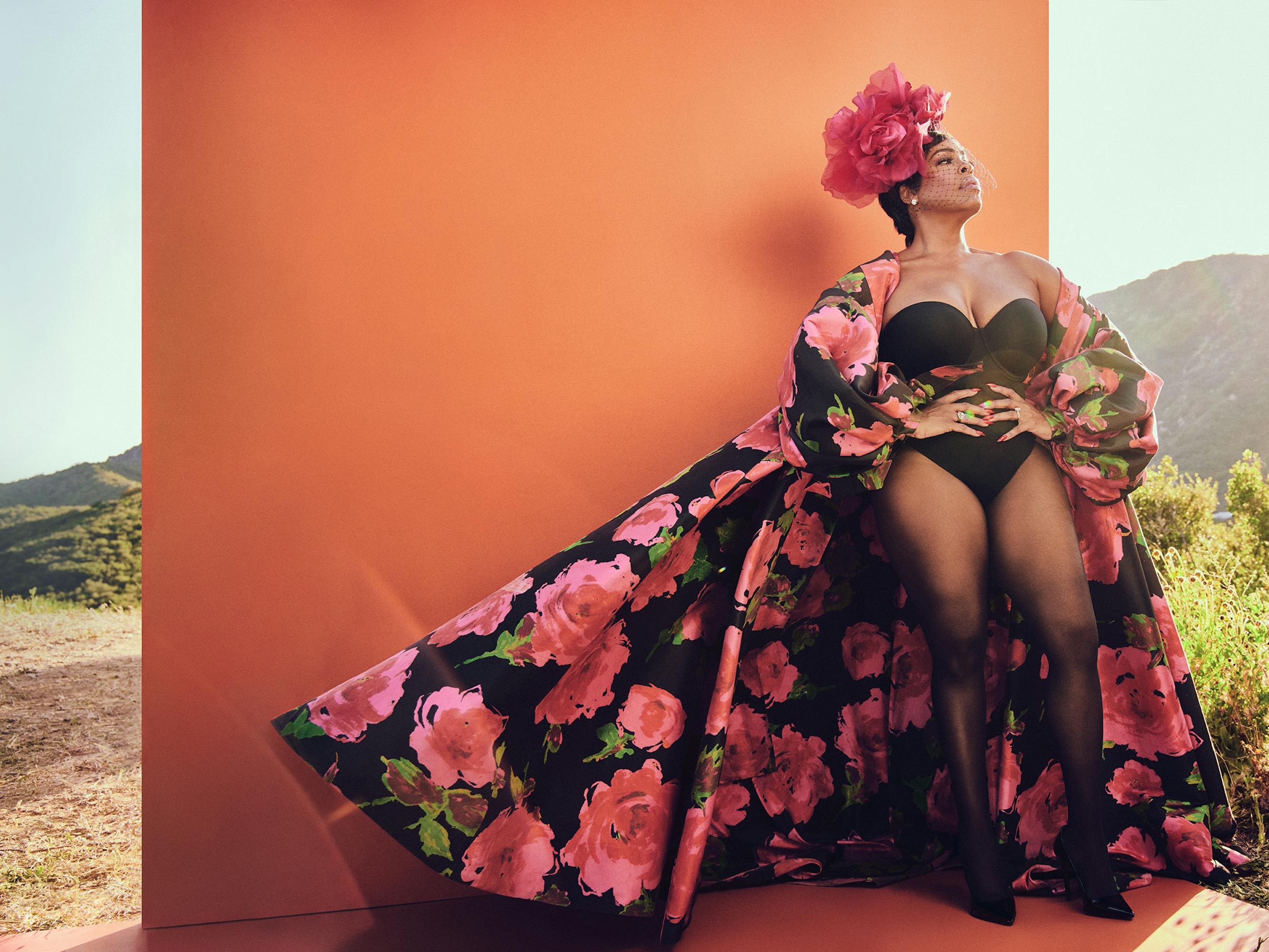 Niecy Nash-Betts strikes a pose against an orange wall. She wears a strapless bodysuit, sheer tights, and a black cape with pink and green flowers on it. Behind the orange wall is a grassy hillside.