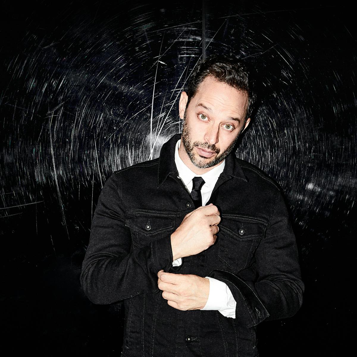Nick Kroll wears a black suit, black tie, and white shirt and looks at the camera. Behind him is a black background with his name written many times in light grey.