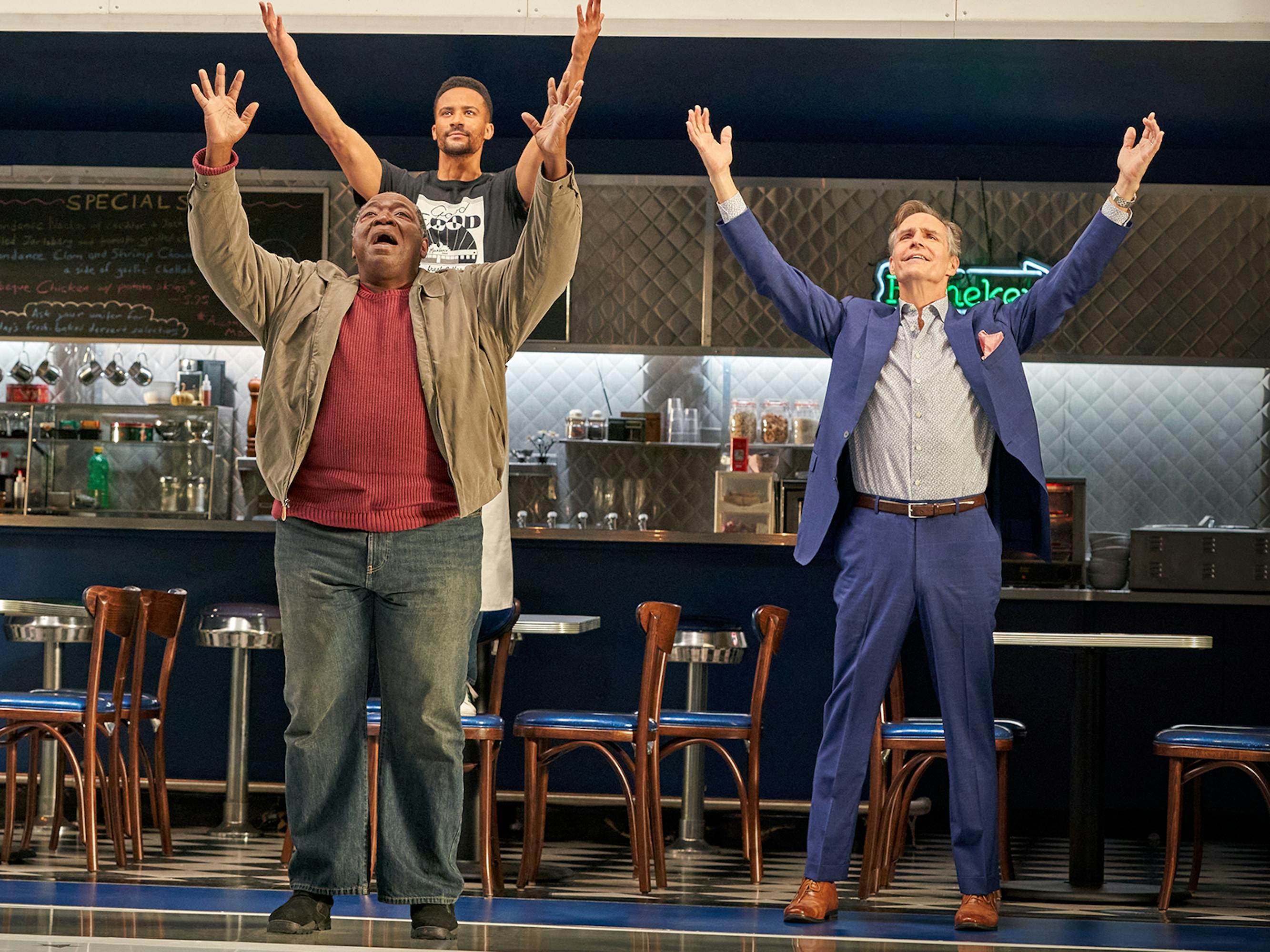 Ben Levi Ross, Chuck Cooper, Austin Goodwin, and Howard McGillin stand outside the Moondance diner with their arms stretched the sky.